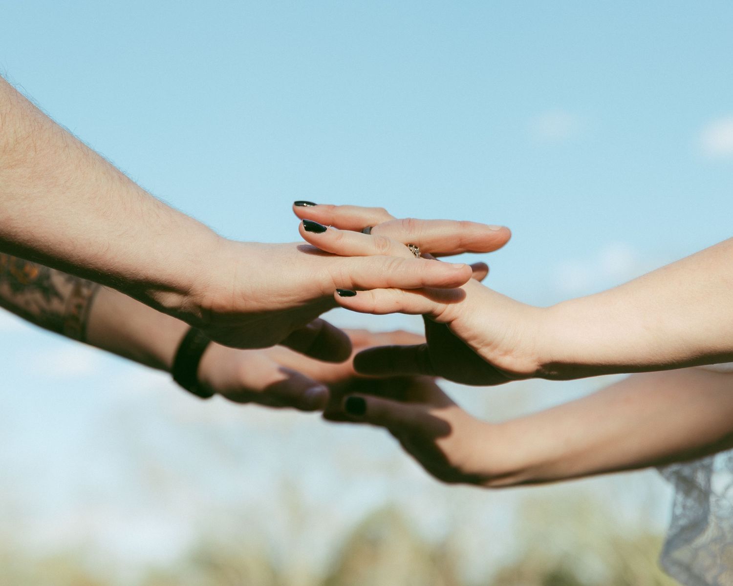 Three people stacking hands together outdoors against a blue sky, symbolizing unity and teamwork.