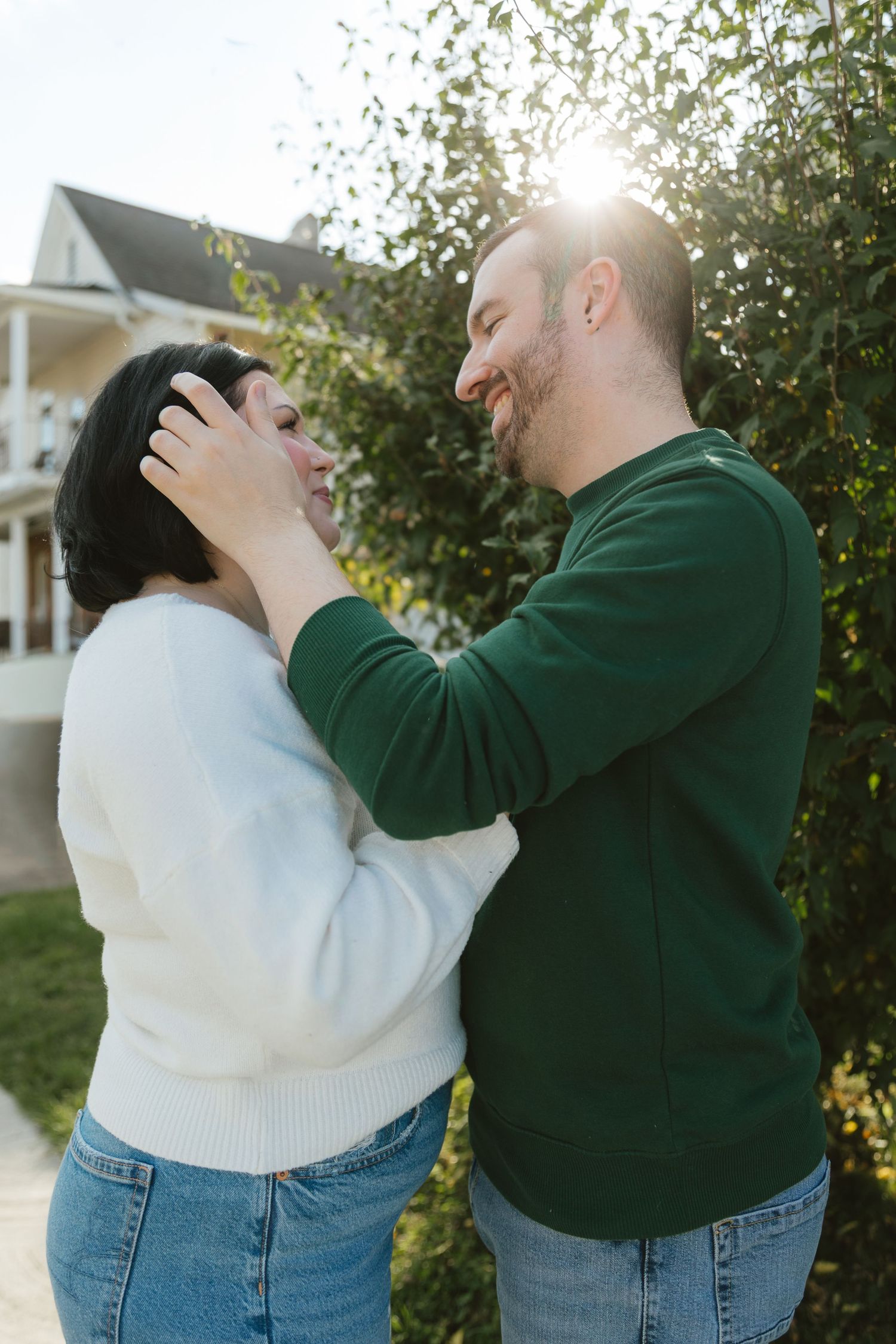 A smiling couple shares a tender moment outdoors, with the man gently touching the woman's face near a green hedge.