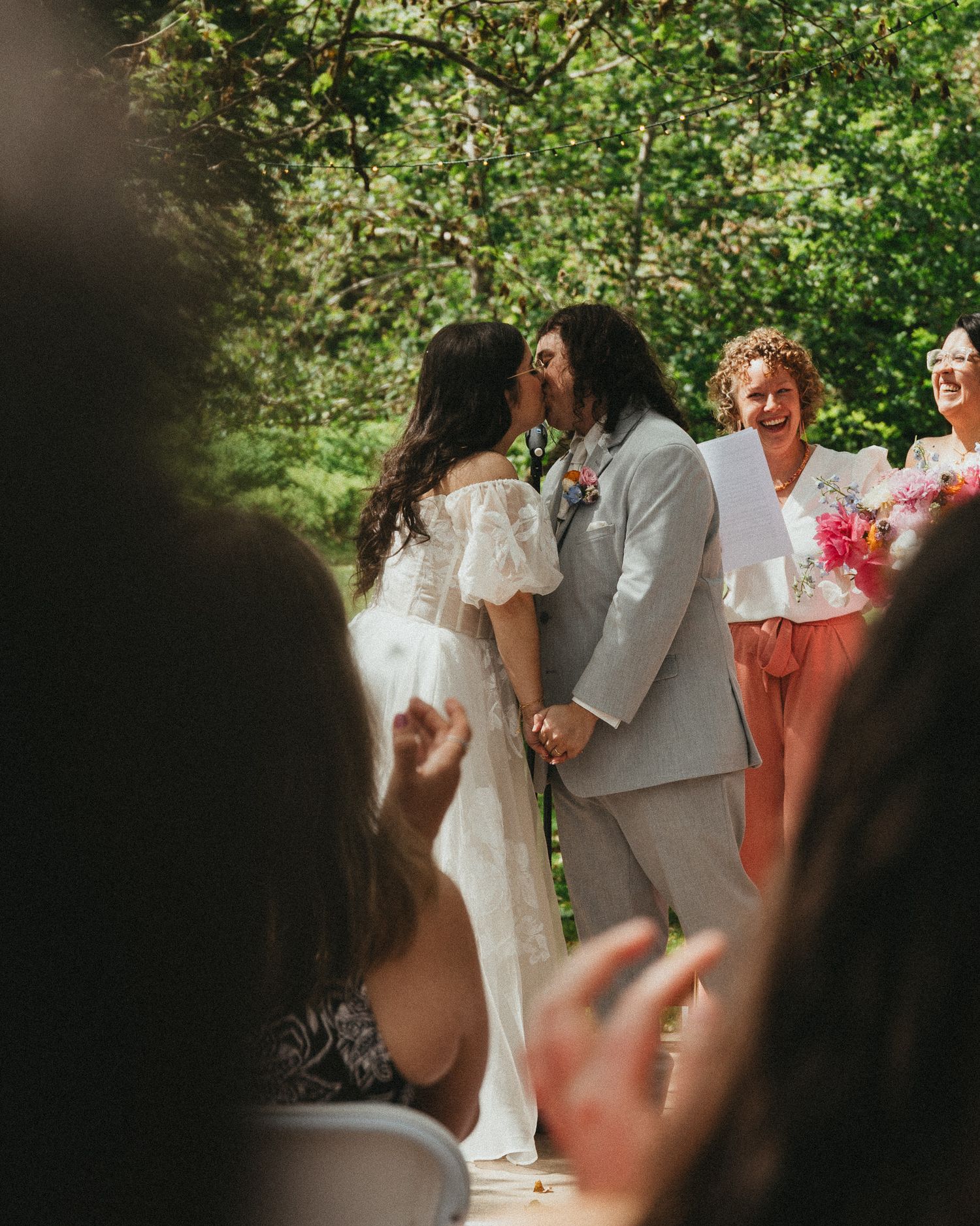 Bride and groom share first kiss at outdoor wedding ceremony while officiant and guests celebrate joyfully.