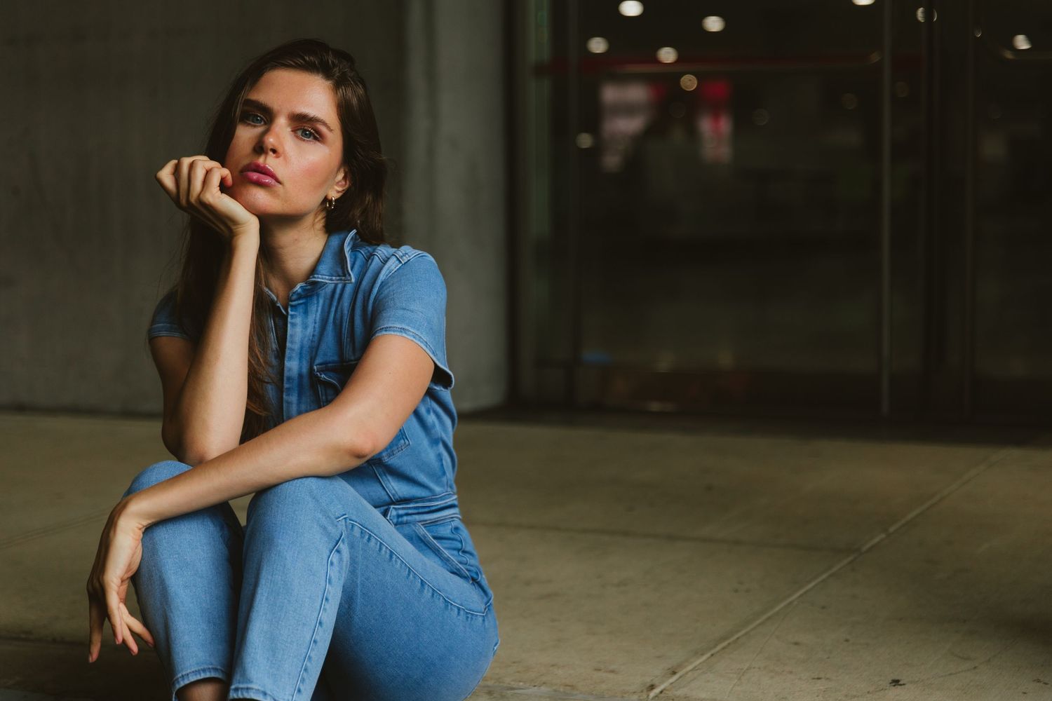 Brunette woman in denim outfit sitting on ground in urban setting.