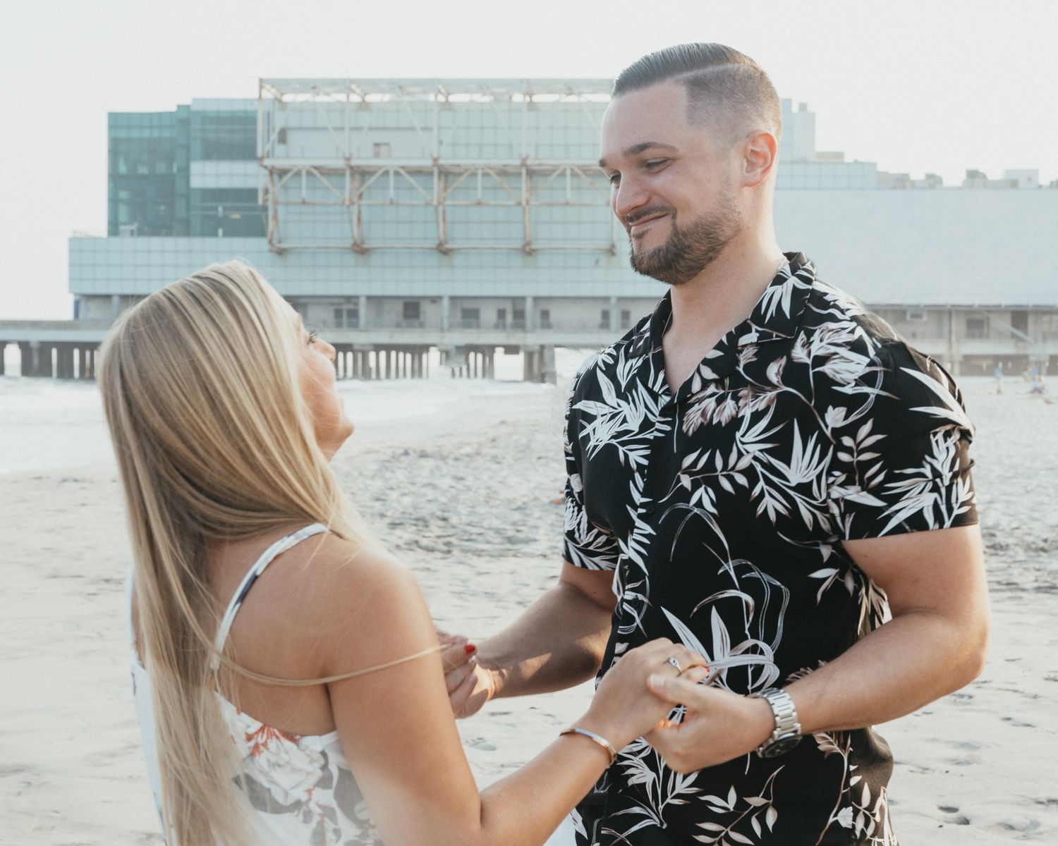 Couple holding hands on beach near pier, man in floral shirt smiling at blonde woman in white dress.