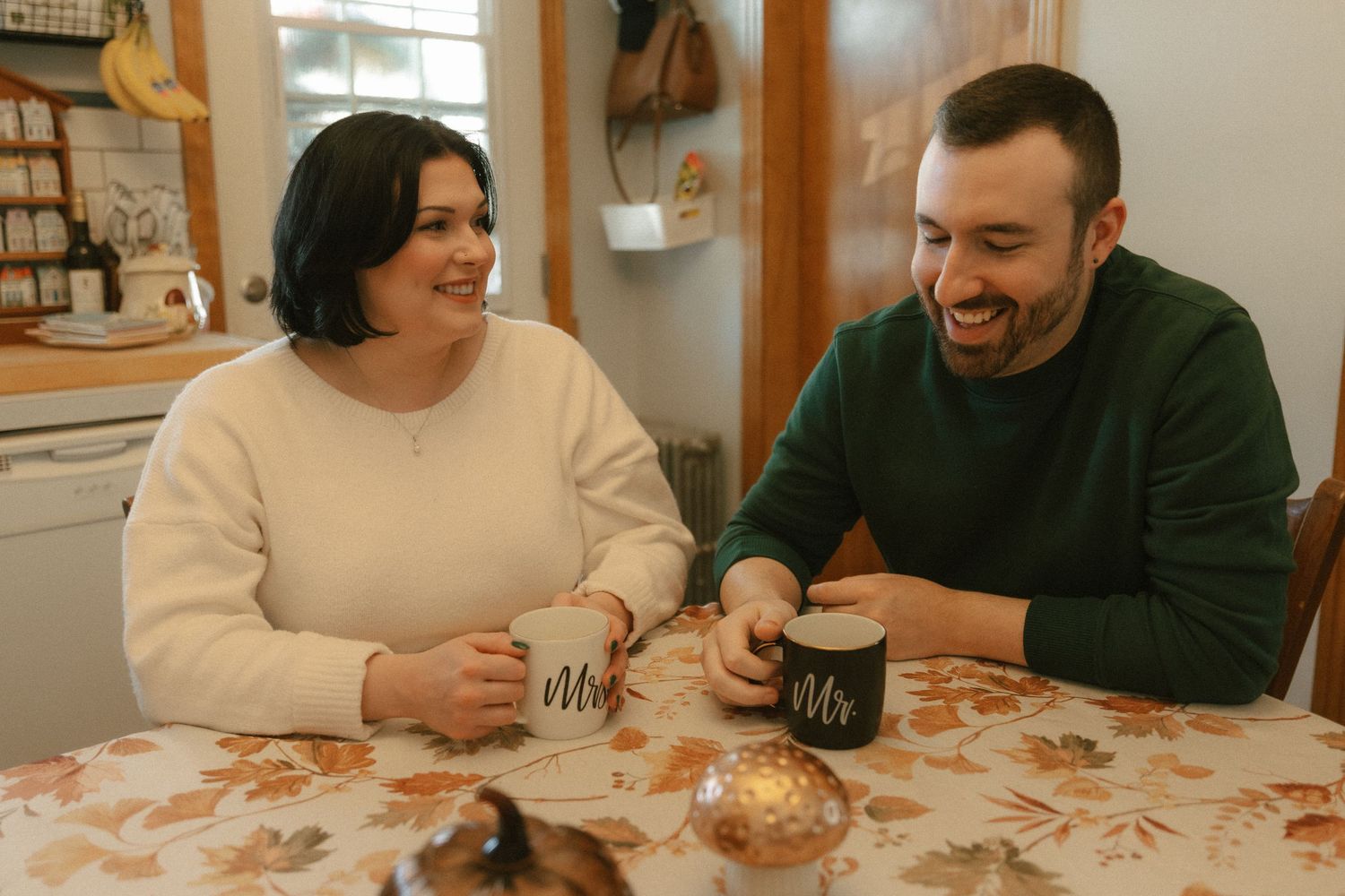 A couple smiles and holds Mr. and Mrs. mugs at a floral tablecloth table in a cozy kitchen setting.