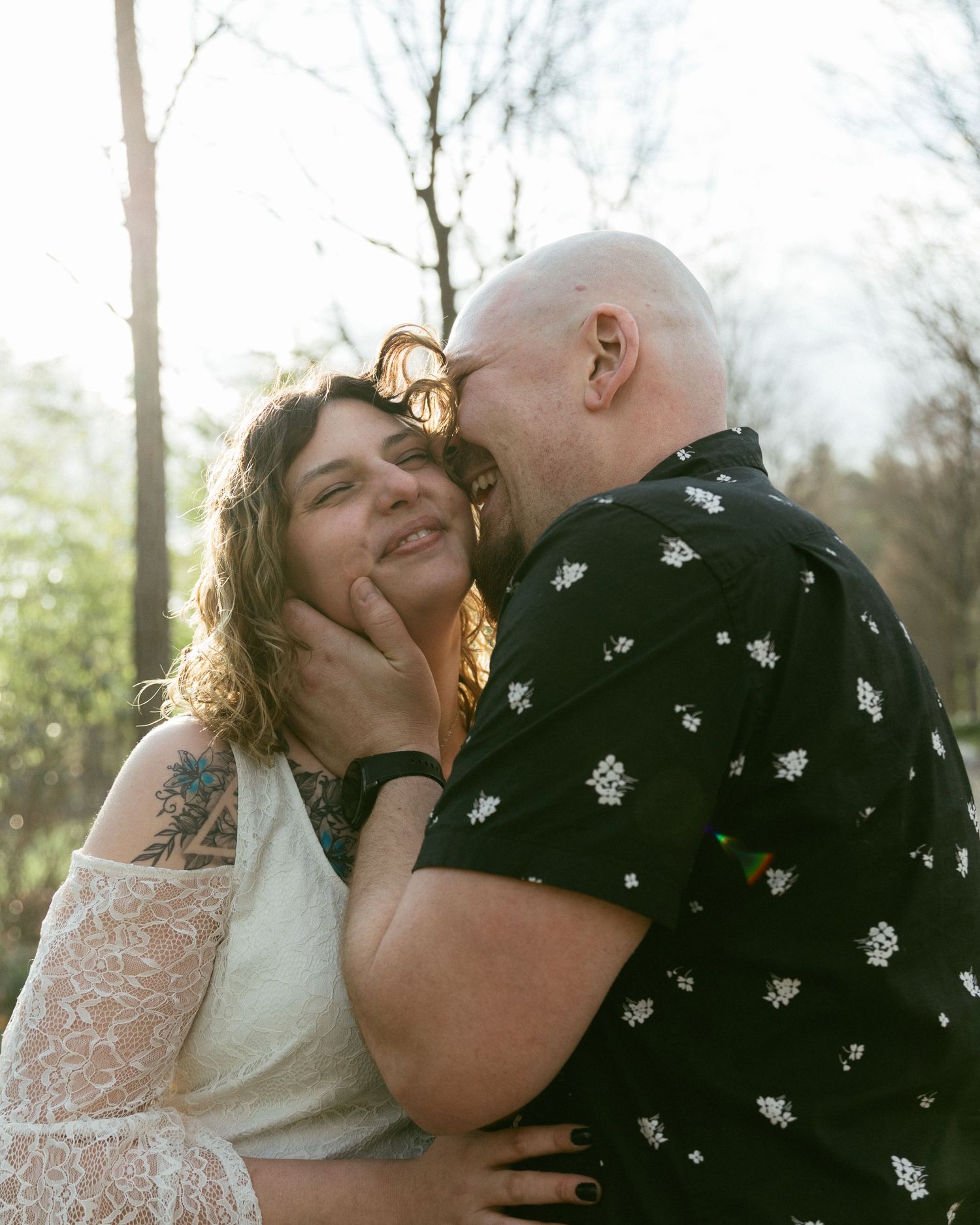 A bald man tenderly kisses a smiling tattooed woman in a lace dress outdoors in golden hour light.