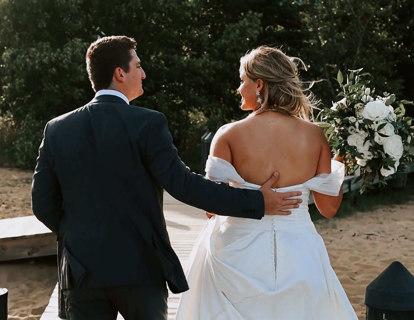 Bride and groom walking together outdoors, bride in off-shoulder white gown holding white floral bouquet.