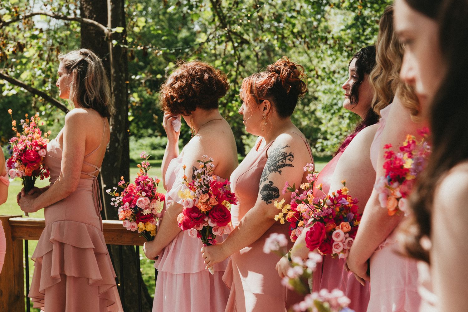 Bridesmaids in pink dresses holding vibrant bouquets of pink and red flowers at outdoor wedding ceremony.