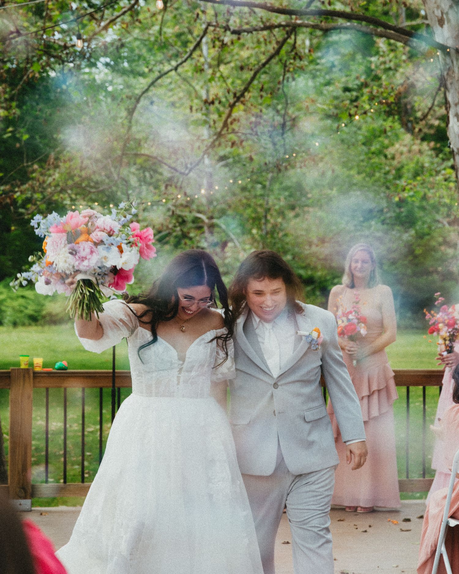 Newlyweds walk down aisle amid colorful smoke, bride raising floral bouquet in celebration at outdoor wedding.