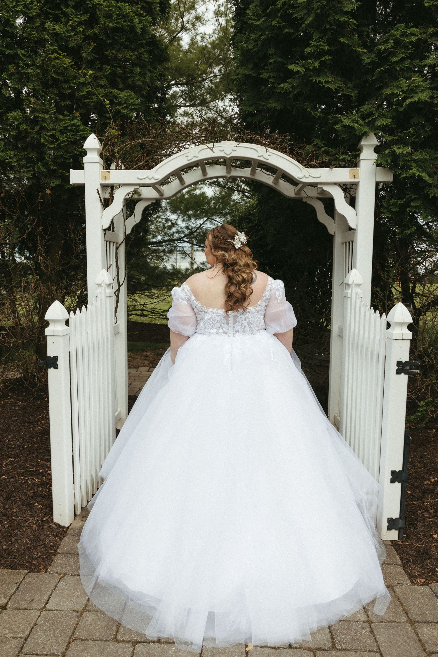 Bride in a white ball gown with lace bodice stands before a white garden arch gate surrounded by lush greenery.