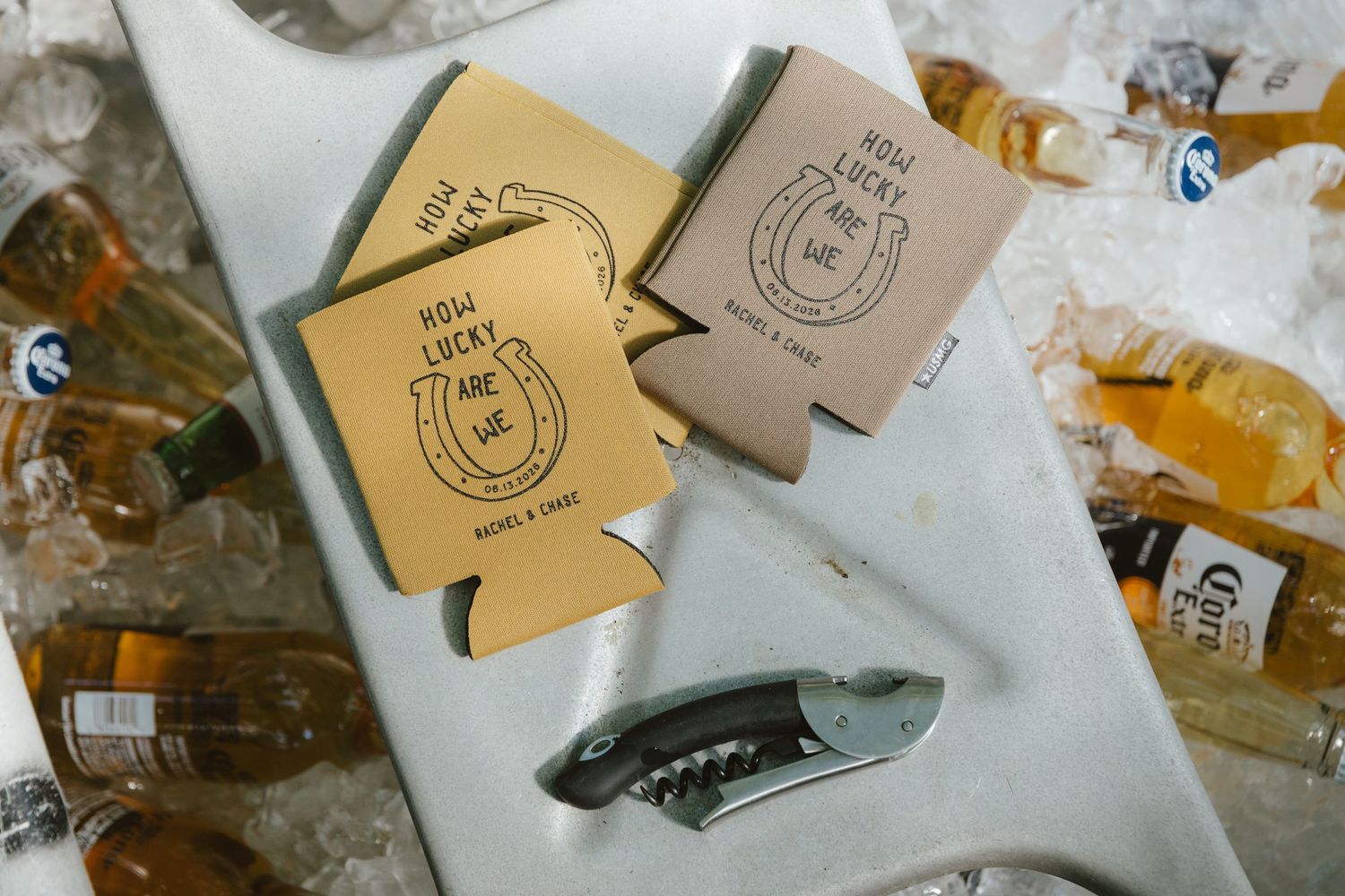 Custom tan koozies reading Now Lucky Are We on ice with Corona bottles and bottle opener at wedding reception.