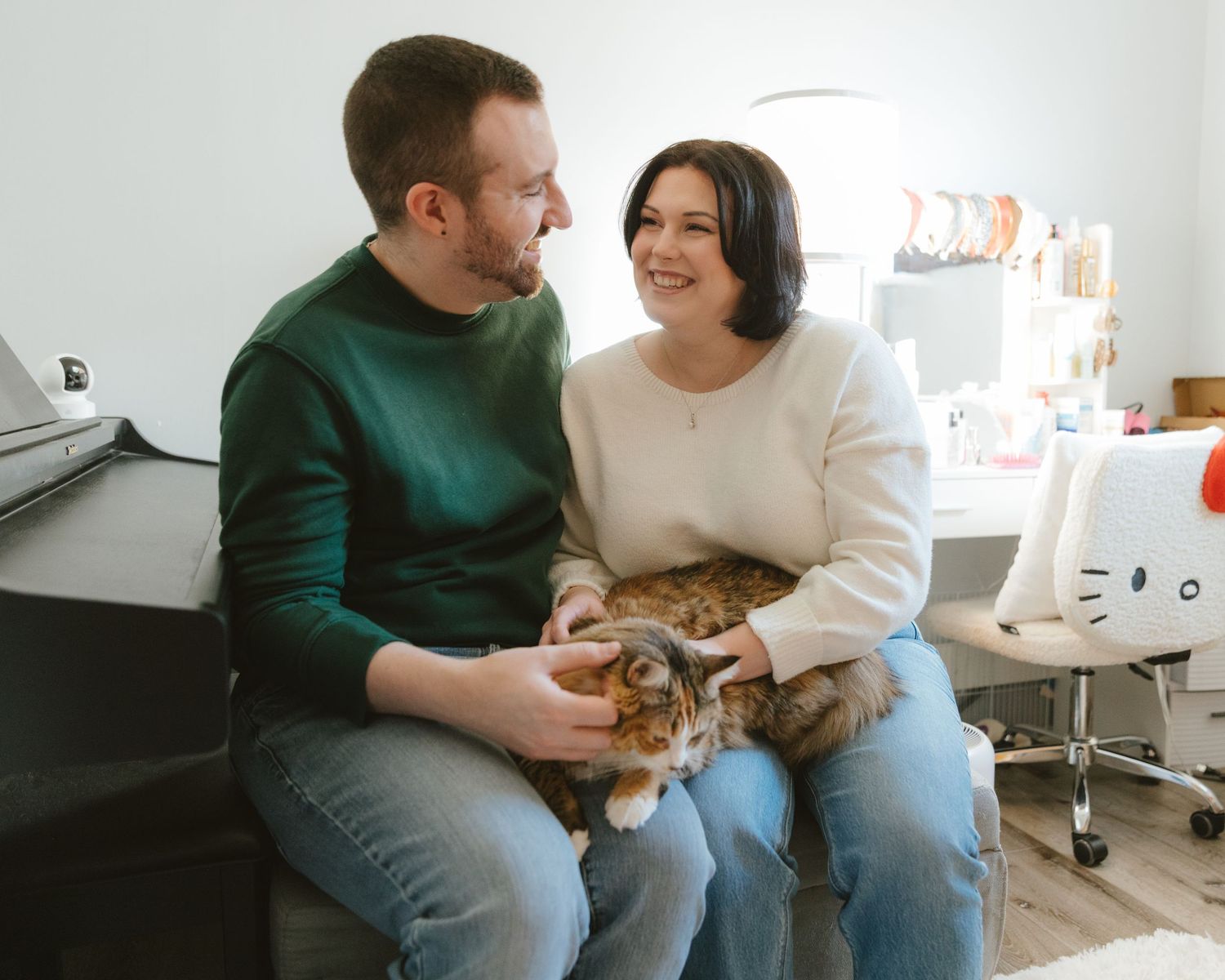 Couple sitting together on a couch smiling, holding a fluffy cat in a bright cozy room.
