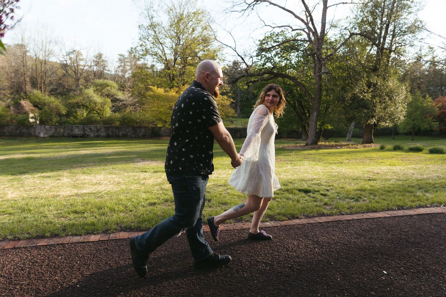 Couple holding hands walking through a lush green park, woman in white dress and man in dark shirt.