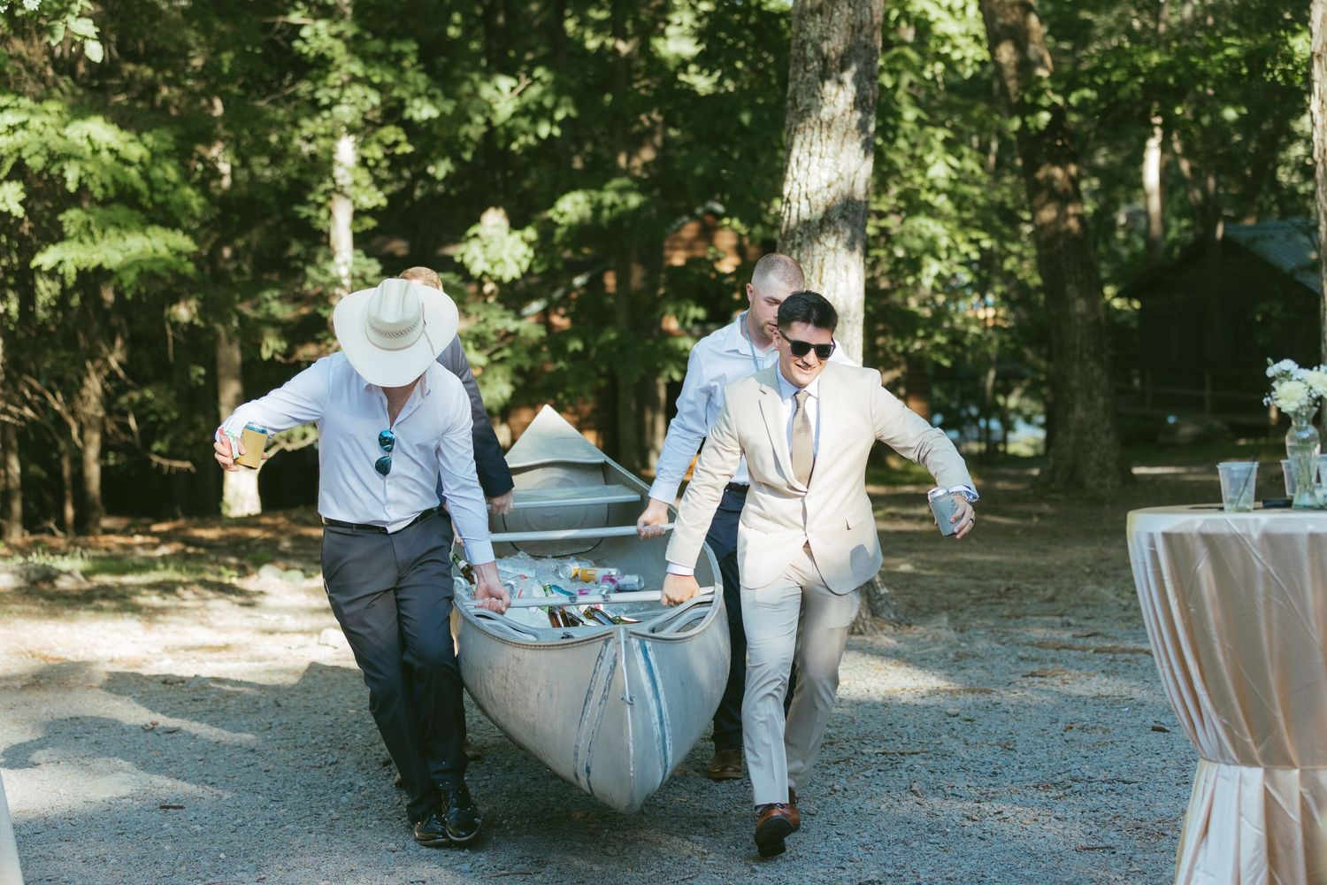 Two men in formal attire carrying a canoe filled with drinks at an outdoor wedding reception.