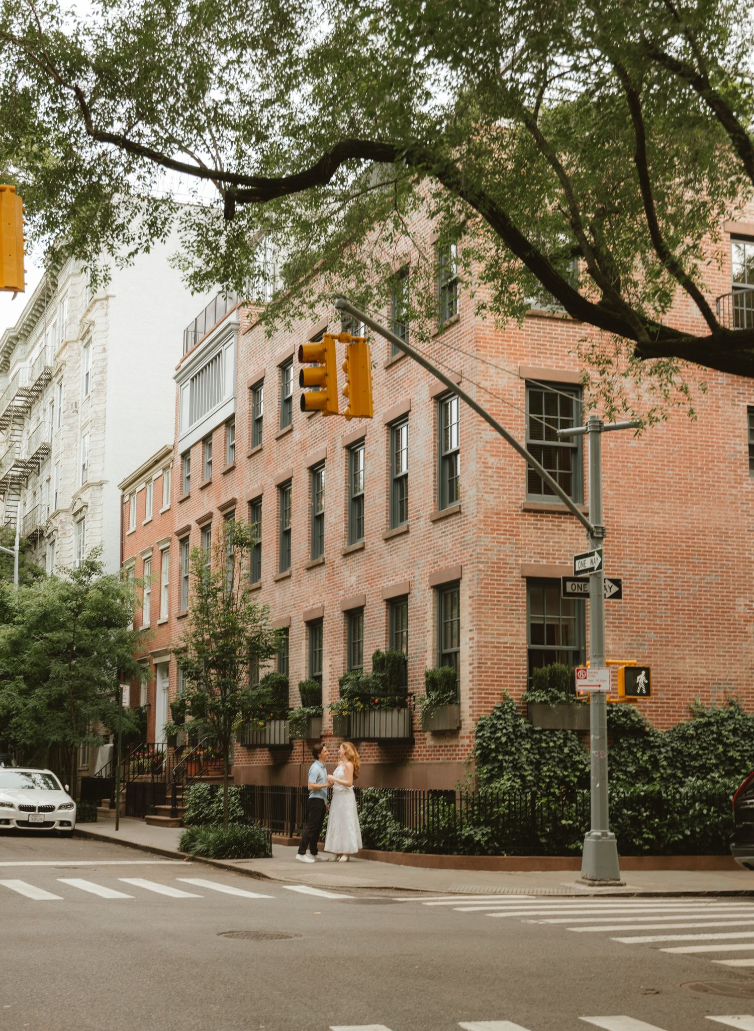 A couple in wedding attire stands at a crosswalk beside a brick building on a tree-lined New York City street.