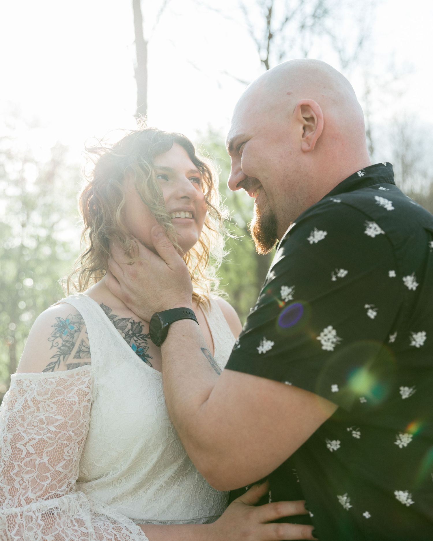 A joyful couple shares a tender moment outdoors, the man cradling the womans face as they laugh together in soft sunlight.