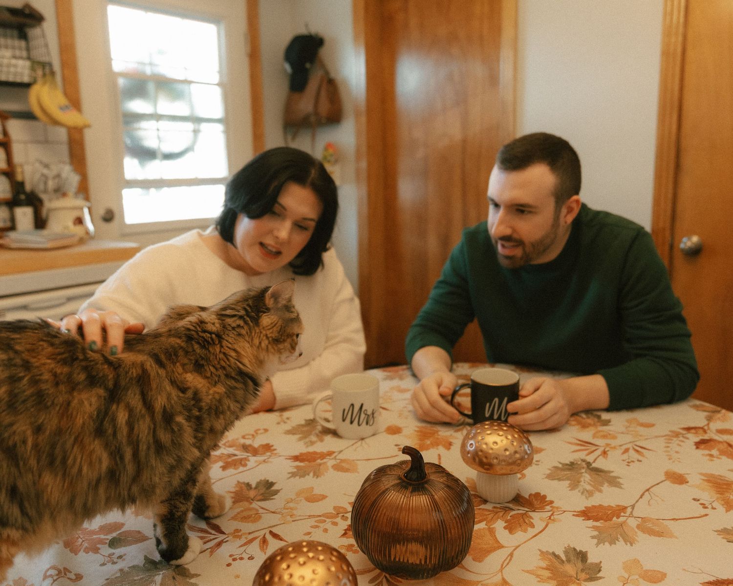 Couple sitting at autumn-decorated table with coffee mugs petting a tabby cat in cozy home kitchen.