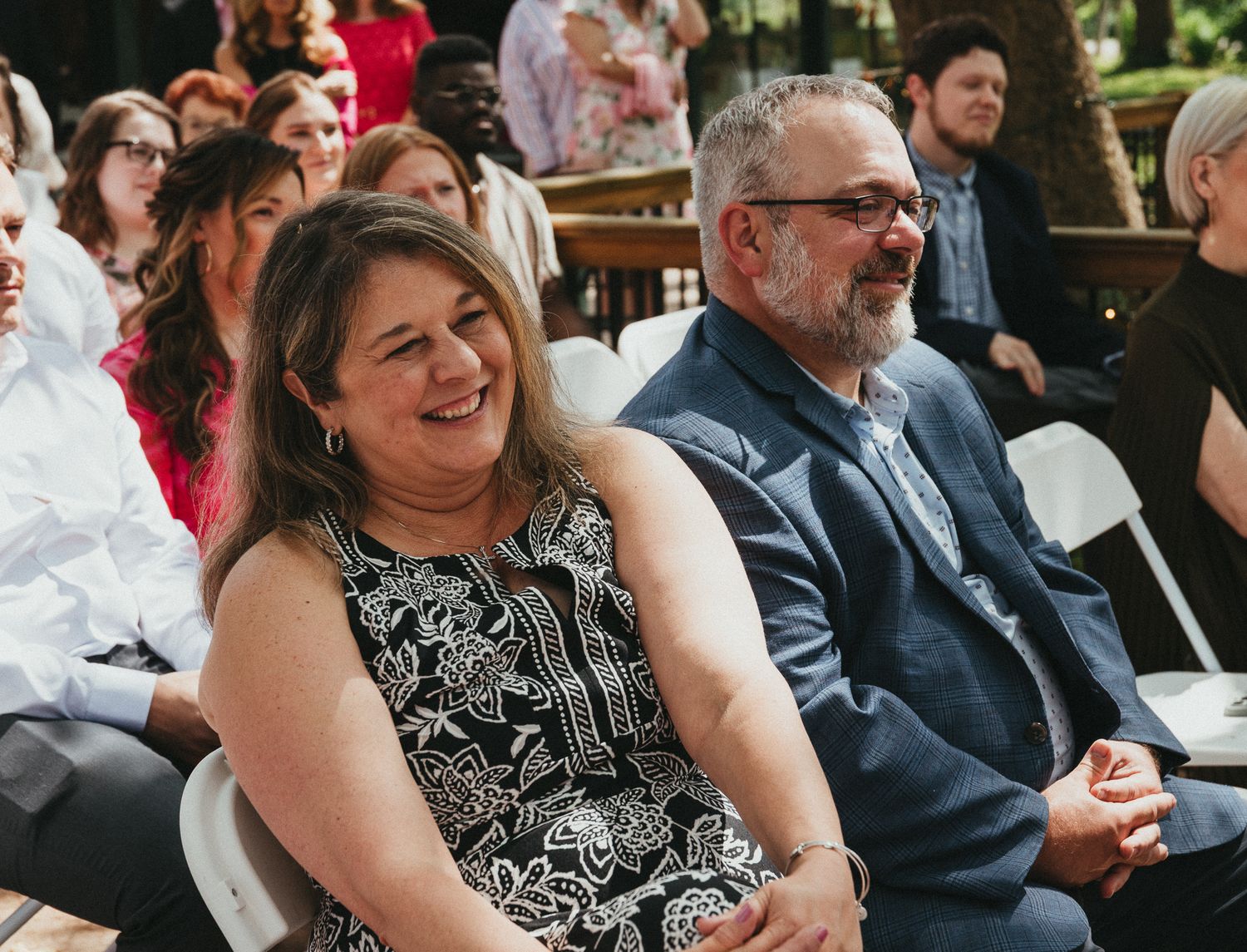 Smiling wedding guests seated at outdoor ceremony, woman in black floral dress and man in navy suit.
