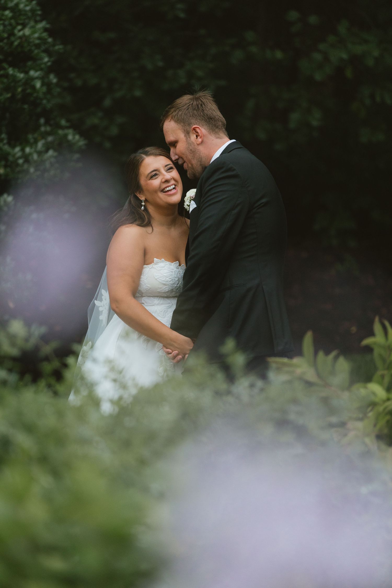 Bride in white strapless gown laughs joyfully as groom in black tuxedo whispers in her ear in a lush garden setting.