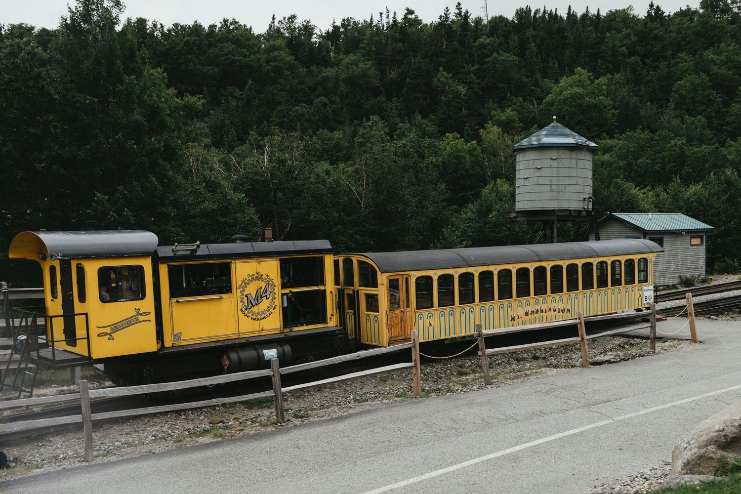 Yellow vintage tourist train parked near a wooden water tower surrounded by green forest.