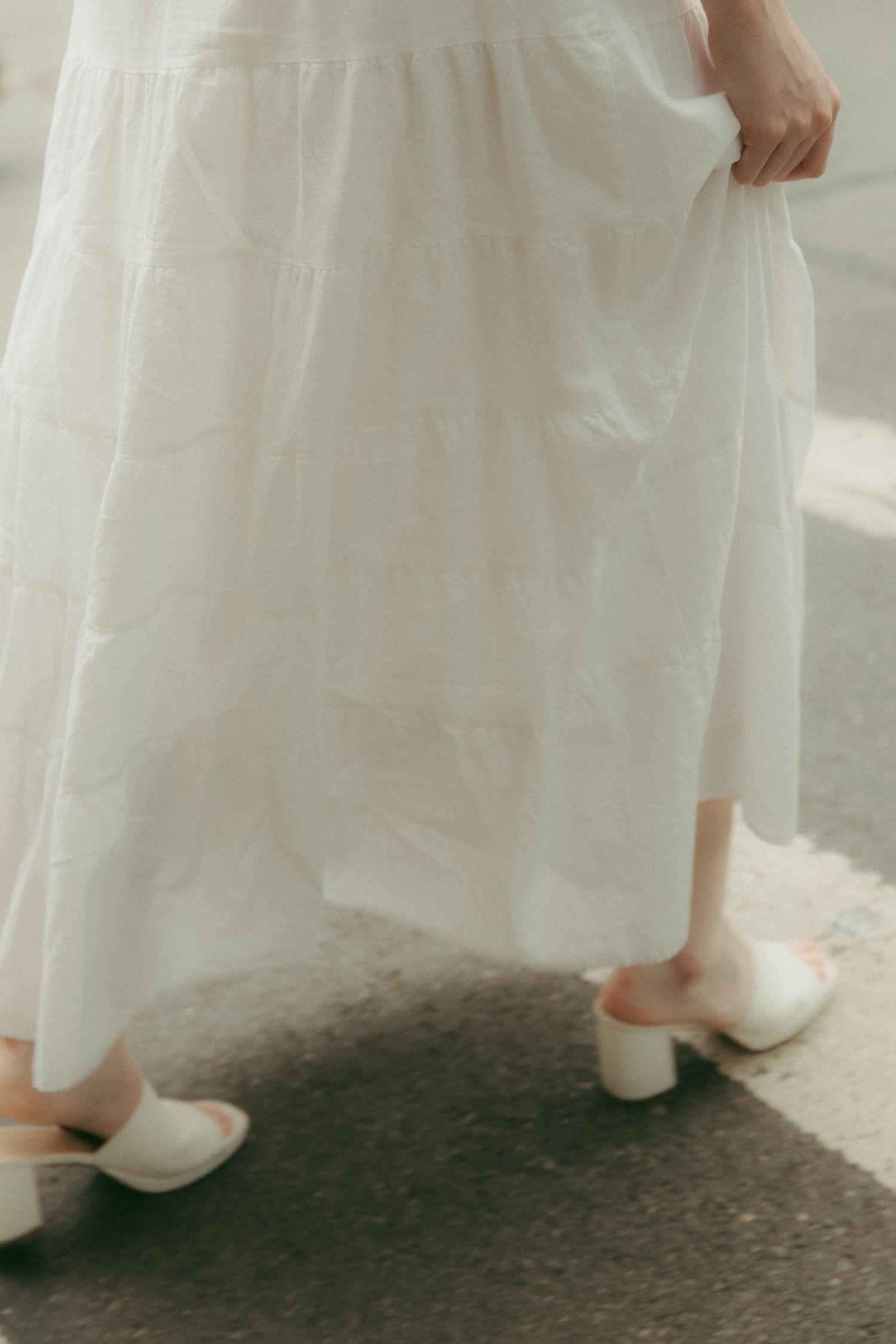 Woman in flowing white tiered maxi dress holding skirt, wearing white block heel mules on pavement.