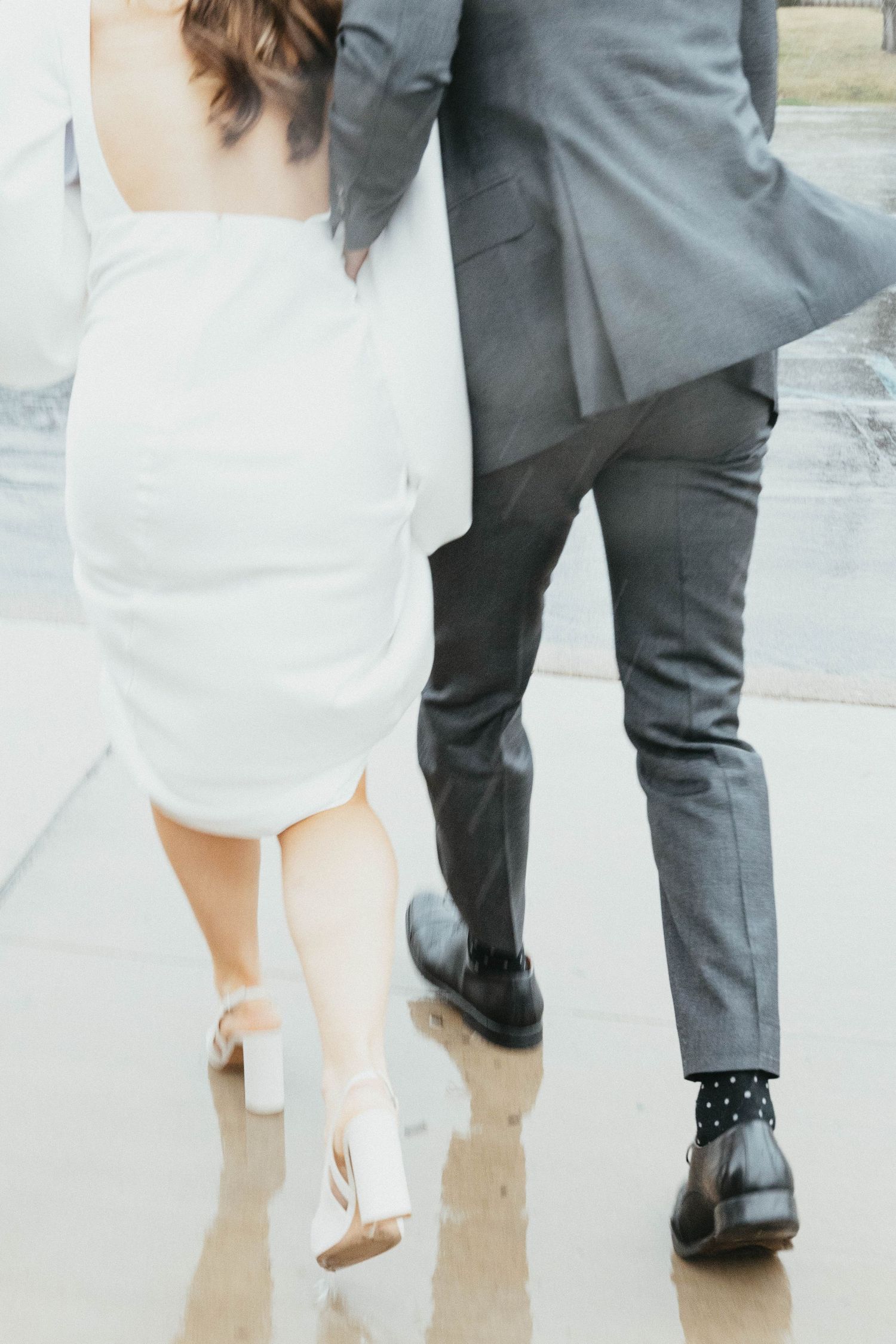 Bride in white mini dress and heels walks with groom in gray suit on wet pavement, shot from behind.
