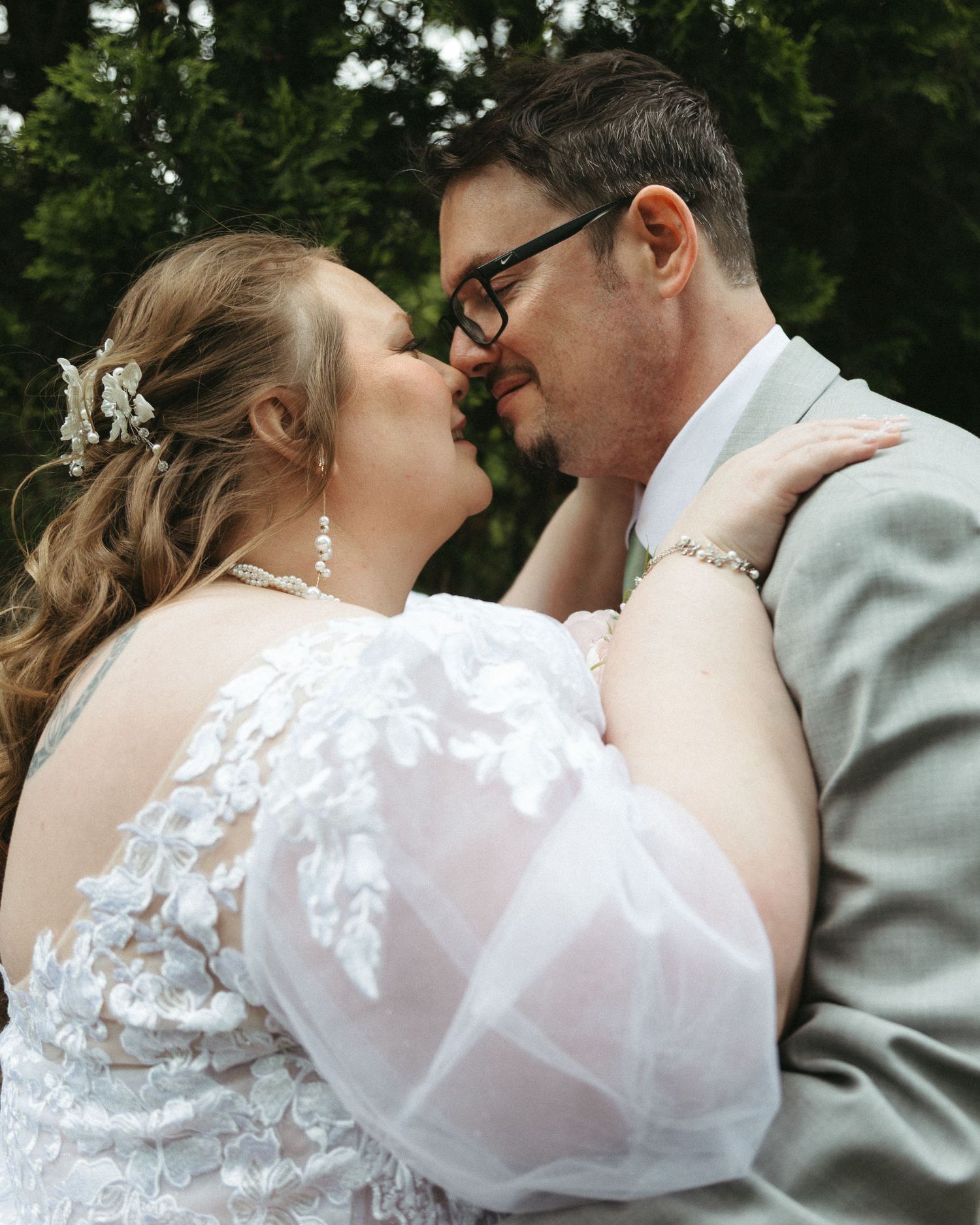 A bride and groom share a romantic kiss outdoors, with the bride in a white lace gown and groom in a gray suit.