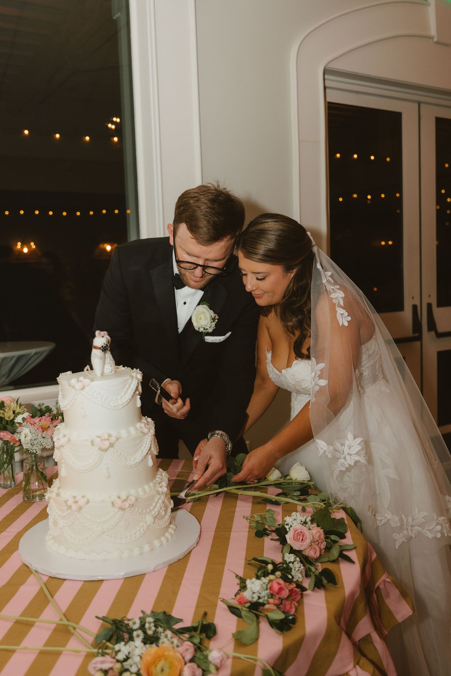 Bride and groom cutting their white three-tier wedding cake at reception table with pink striped tablecloth and floral decor.