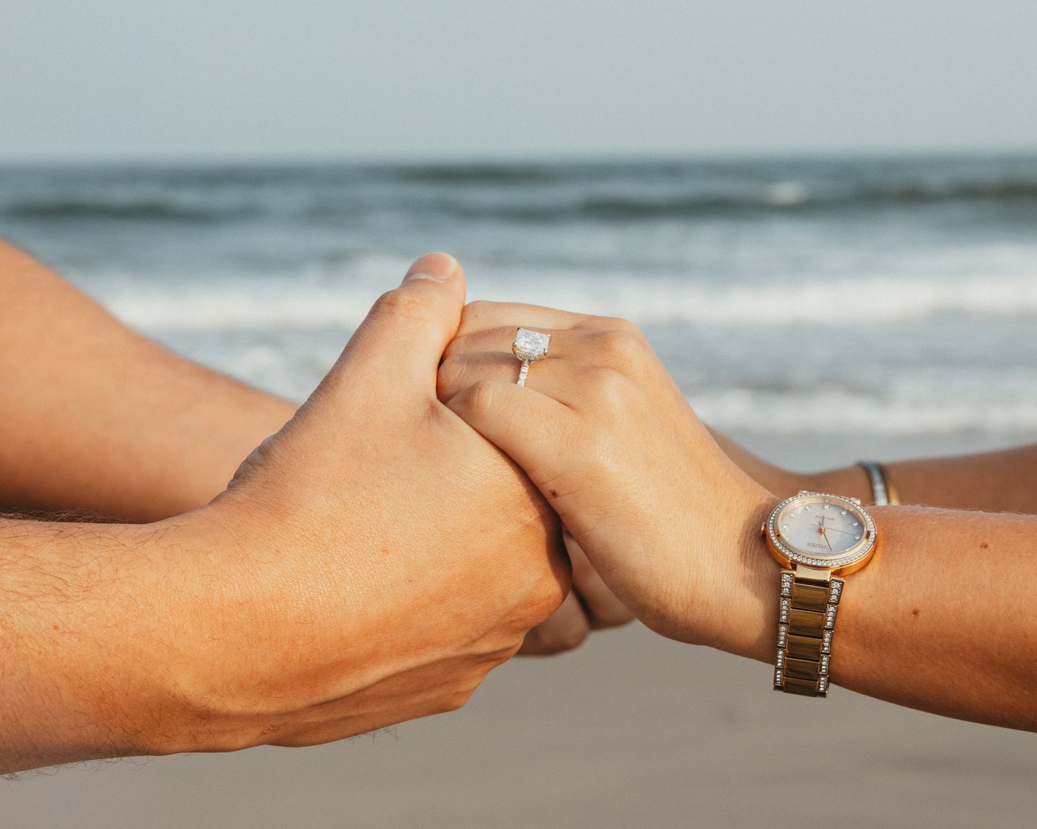 Couple holding hands on beach, woman wearing diamond engagement ring and gold watch by the ocean.
