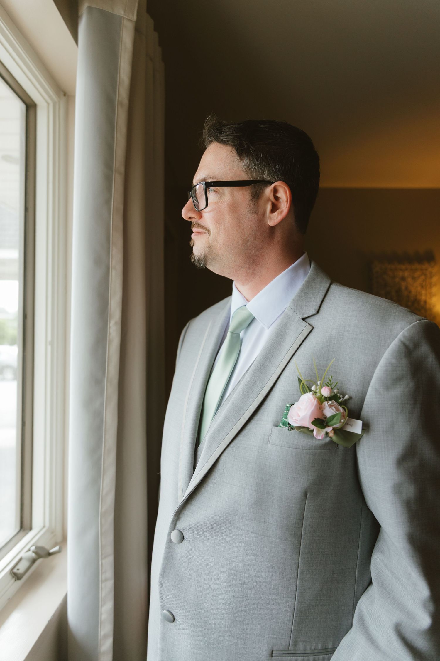 Groom in light gray suit with mint tie and floral boutonniere gazes out window on wedding day.