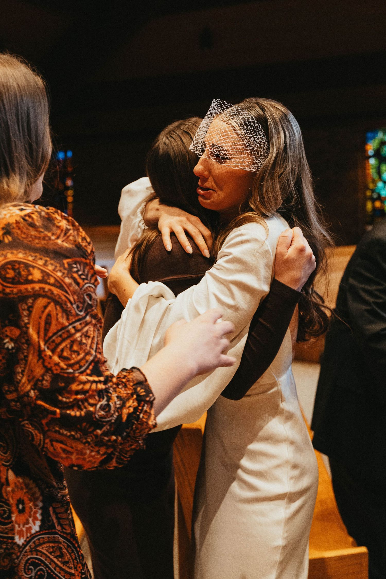 Bride in white gown and birdcage veil shares emotional hug with guest inside a warmly lit church.
