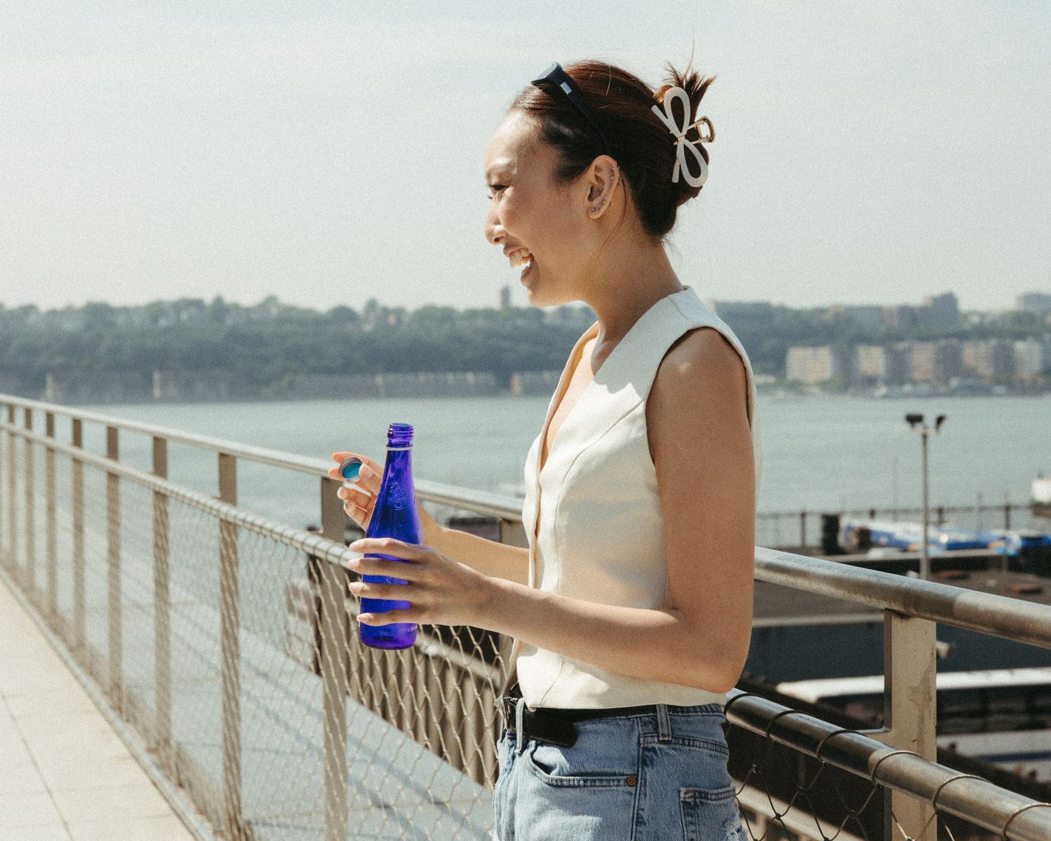 Woman holding blue bottle on waterfront bridge on sunny day.