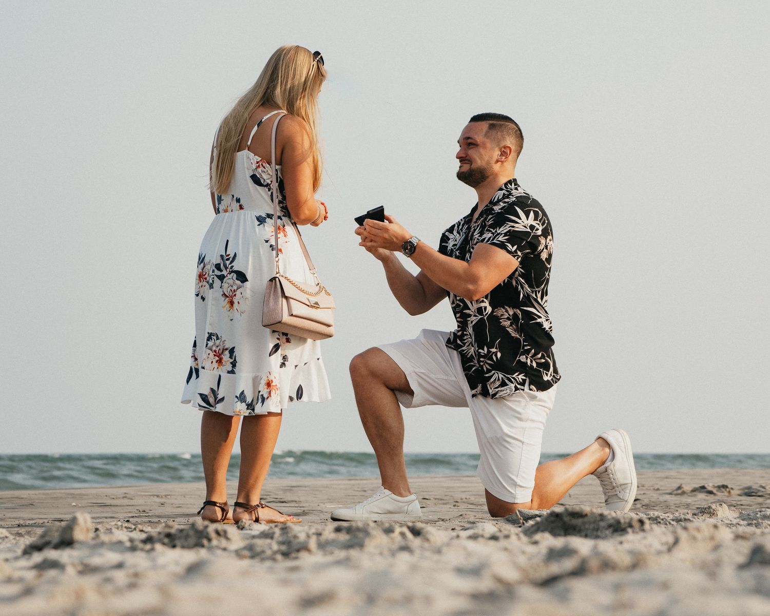 Man proposing on one knee to woman on sandy beach holding ring box near ocean shore.