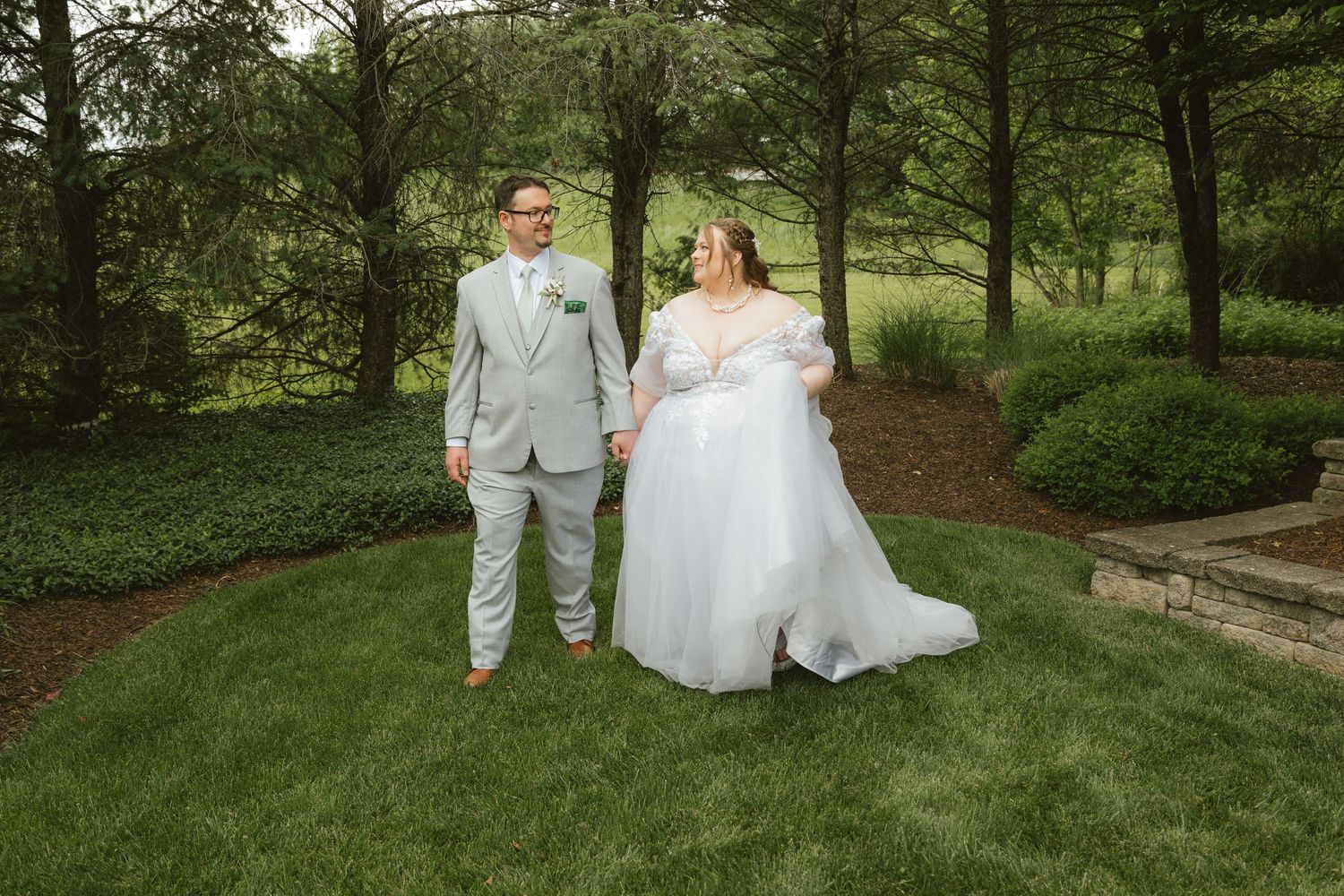 A bride in a white off-shoulder gown and groom in a gray suit hold hands outdoors on lush green grass.