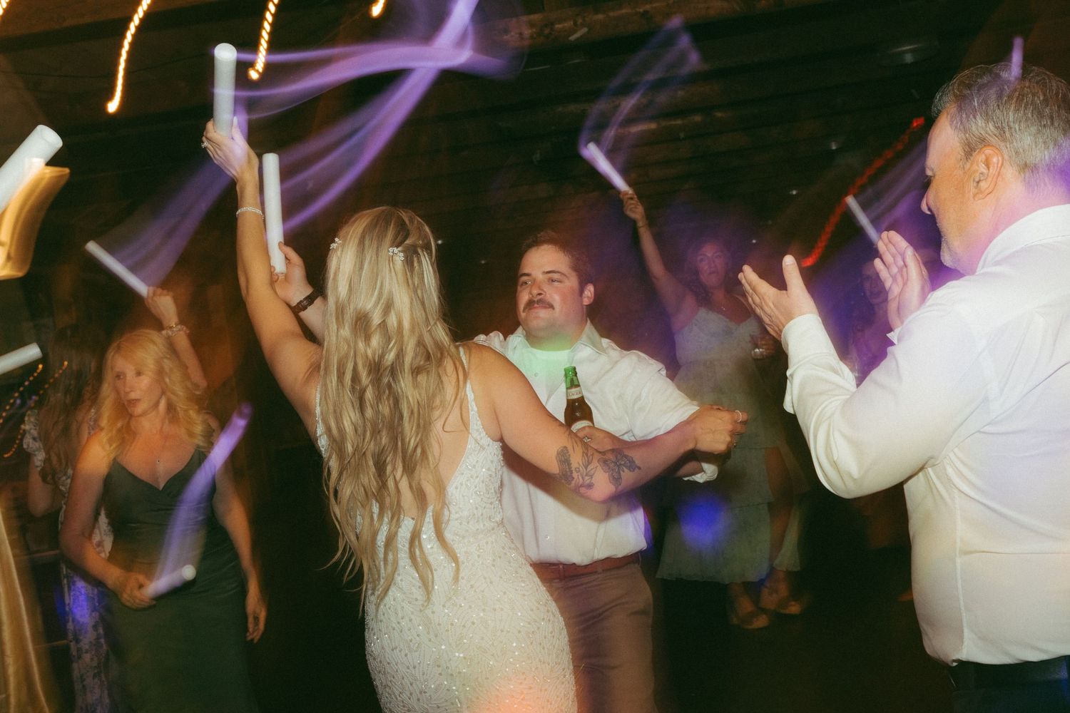Bride and groom dancing with guests holding glow sticks at a lively wedding reception party.