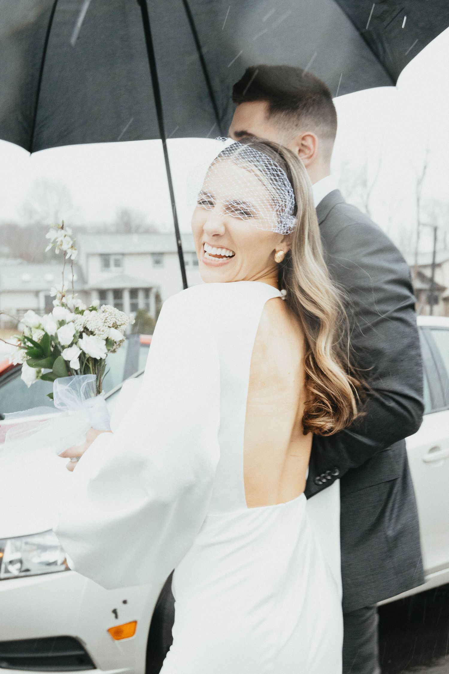 Smiling bride in backless white gown holds bouquet under umbrella with groom on rainy wedding day.