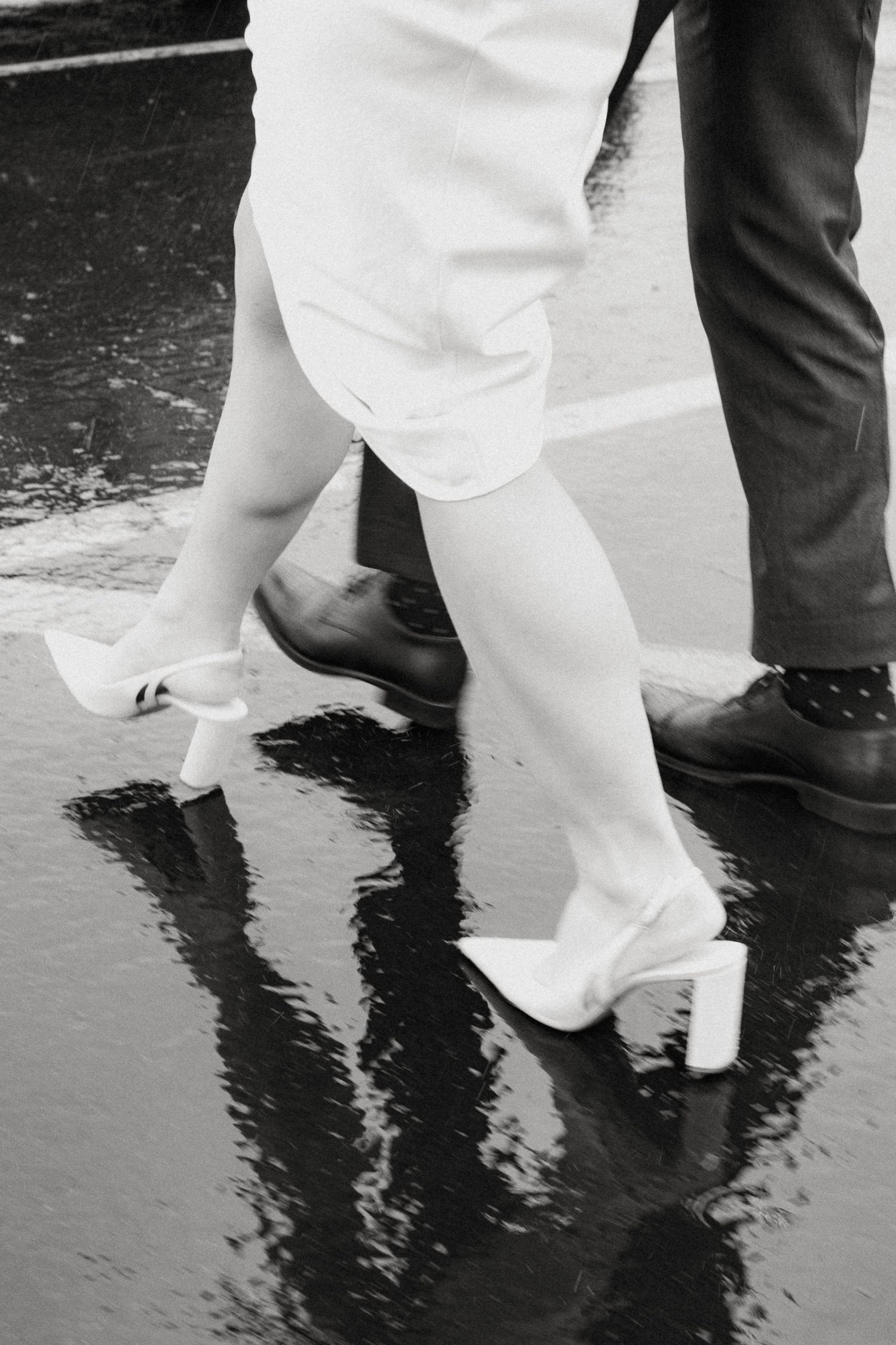 Black and white photo of bride in white heels and groom walking on rain-soaked pavement.