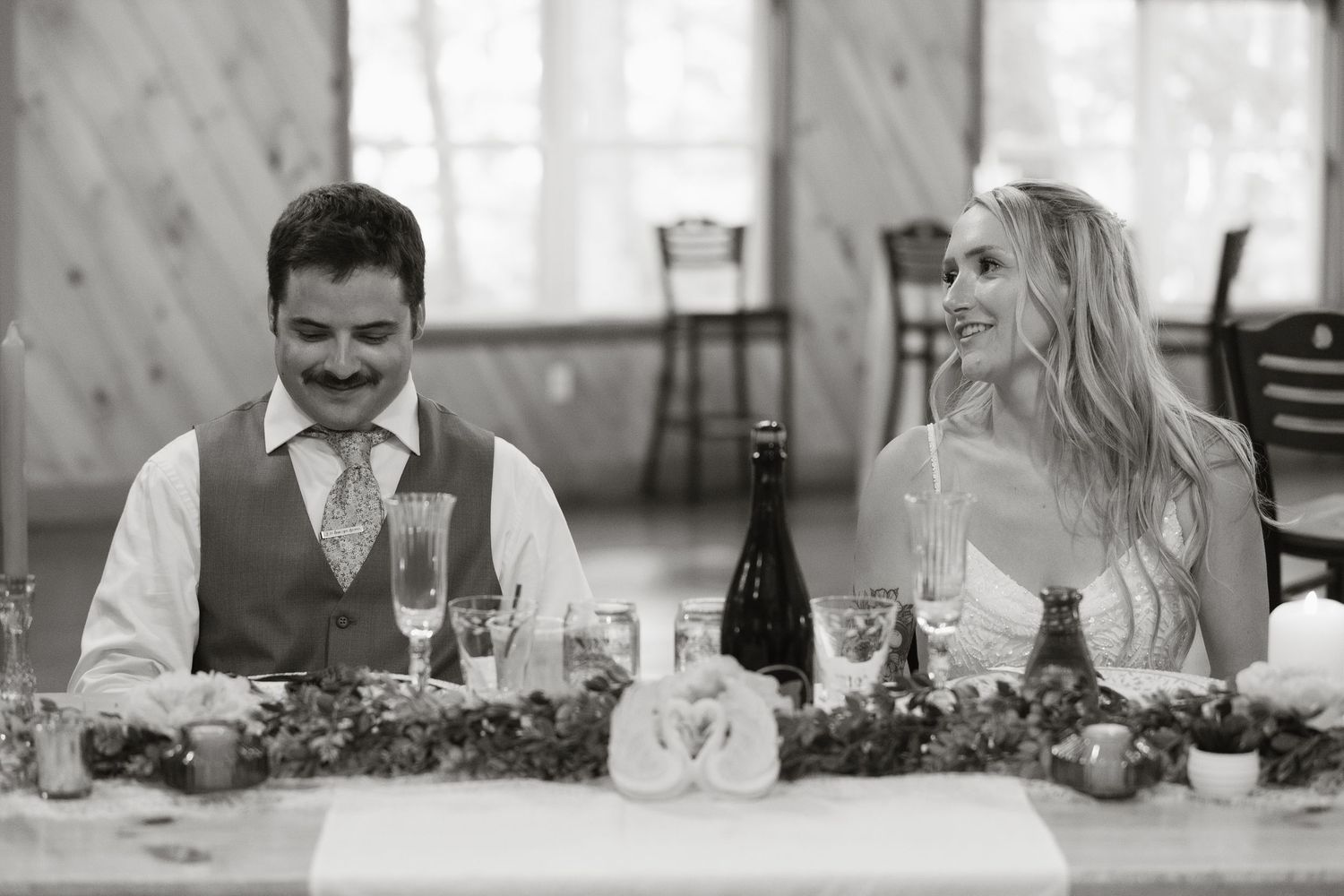 Black and white photo of smiling bride and groom seated at wedding reception sweetheart table with greenery garland.