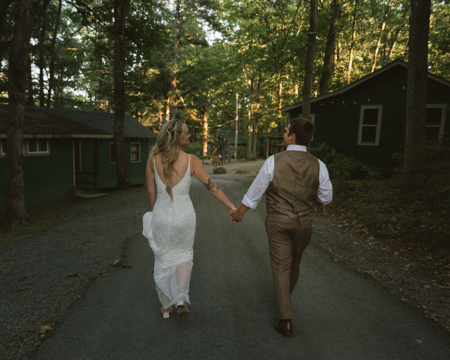 Bride and groom walking hand in hand down a tree-lined path surrounded by rustic cabins in a forest setting.