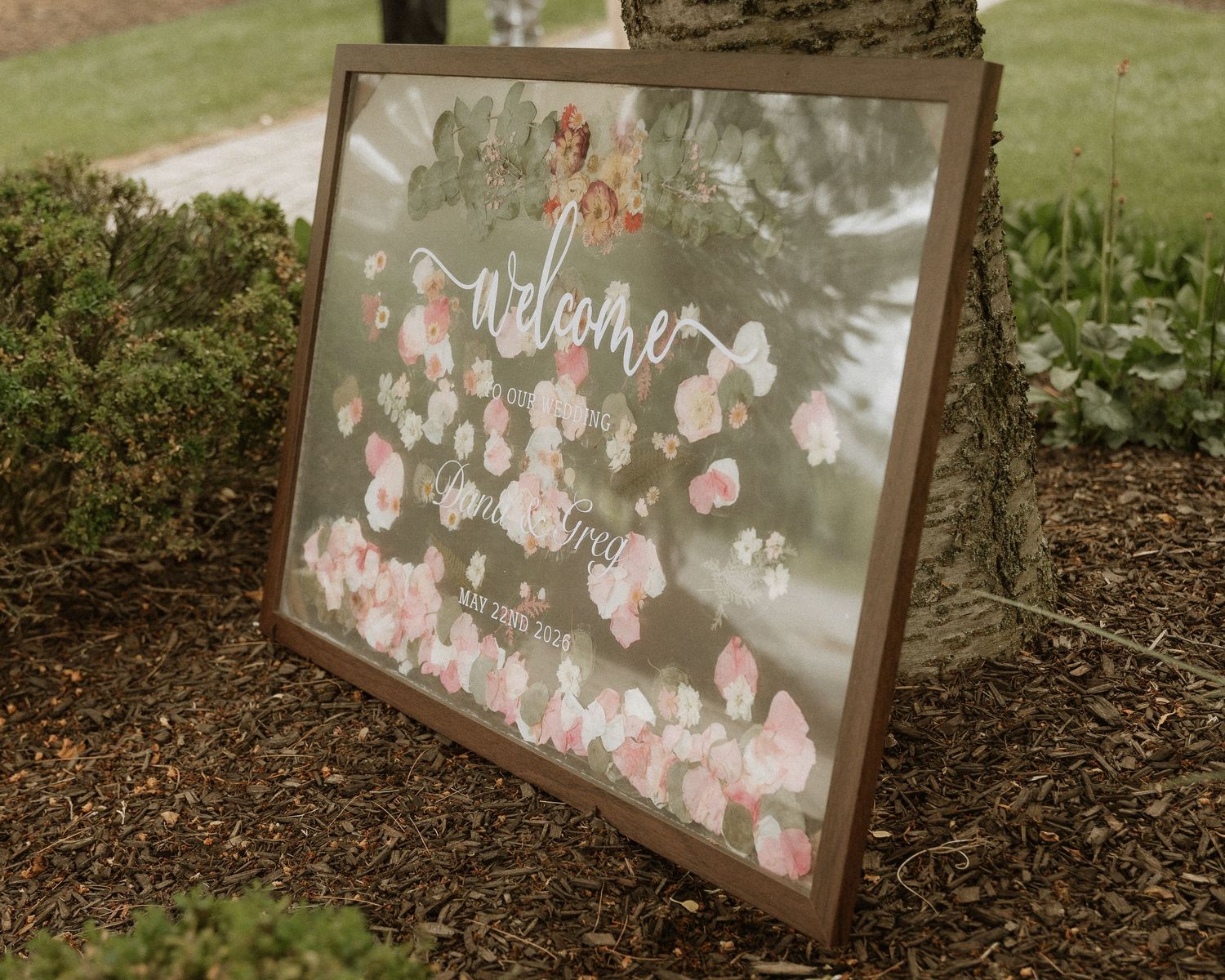 Elegant floral wedding welcome sign with pink roses on glass, framed in wood, leaning against a tree outdoors.