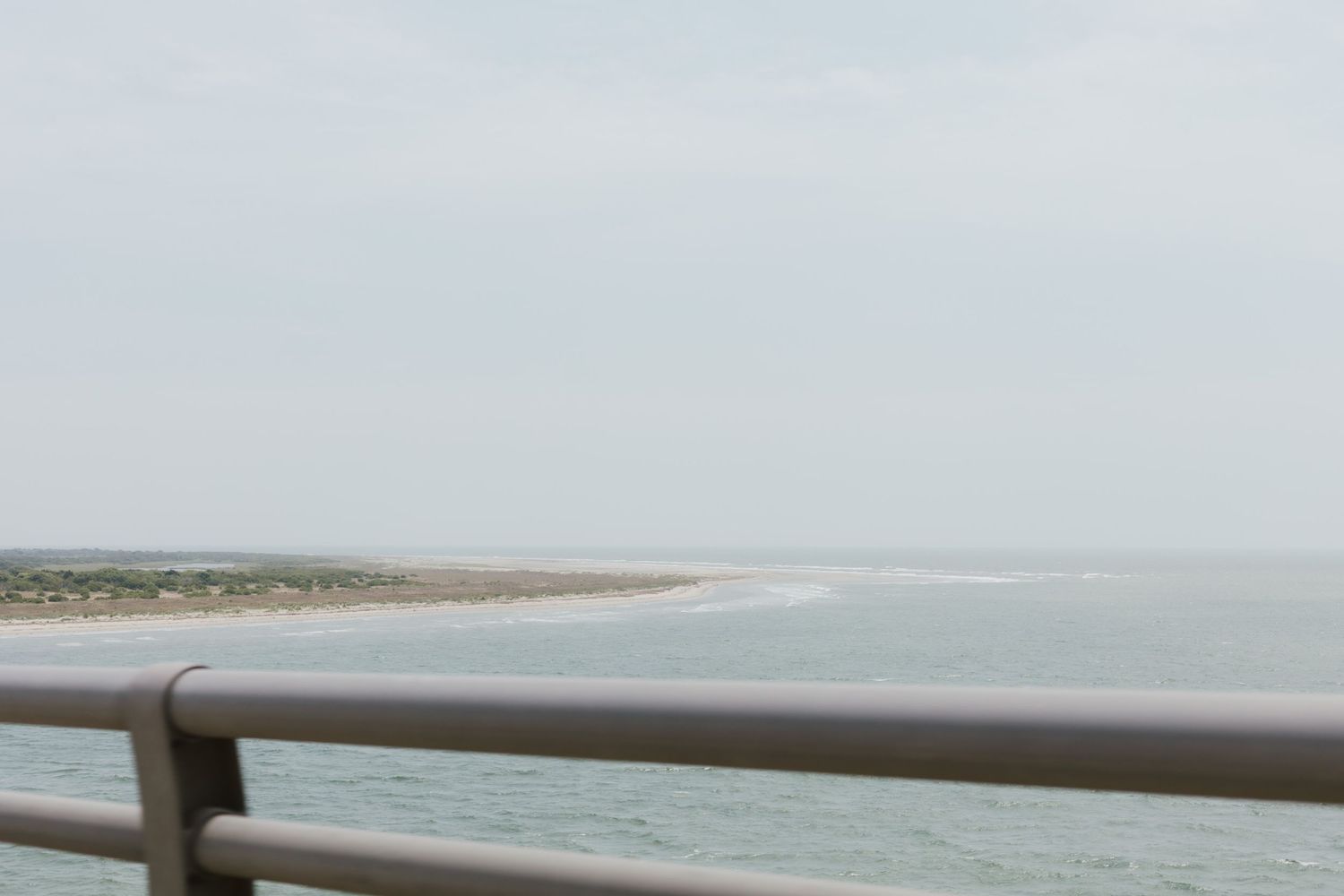 Calm ocean shoreline viewed from a bridge railing on an overcast day.