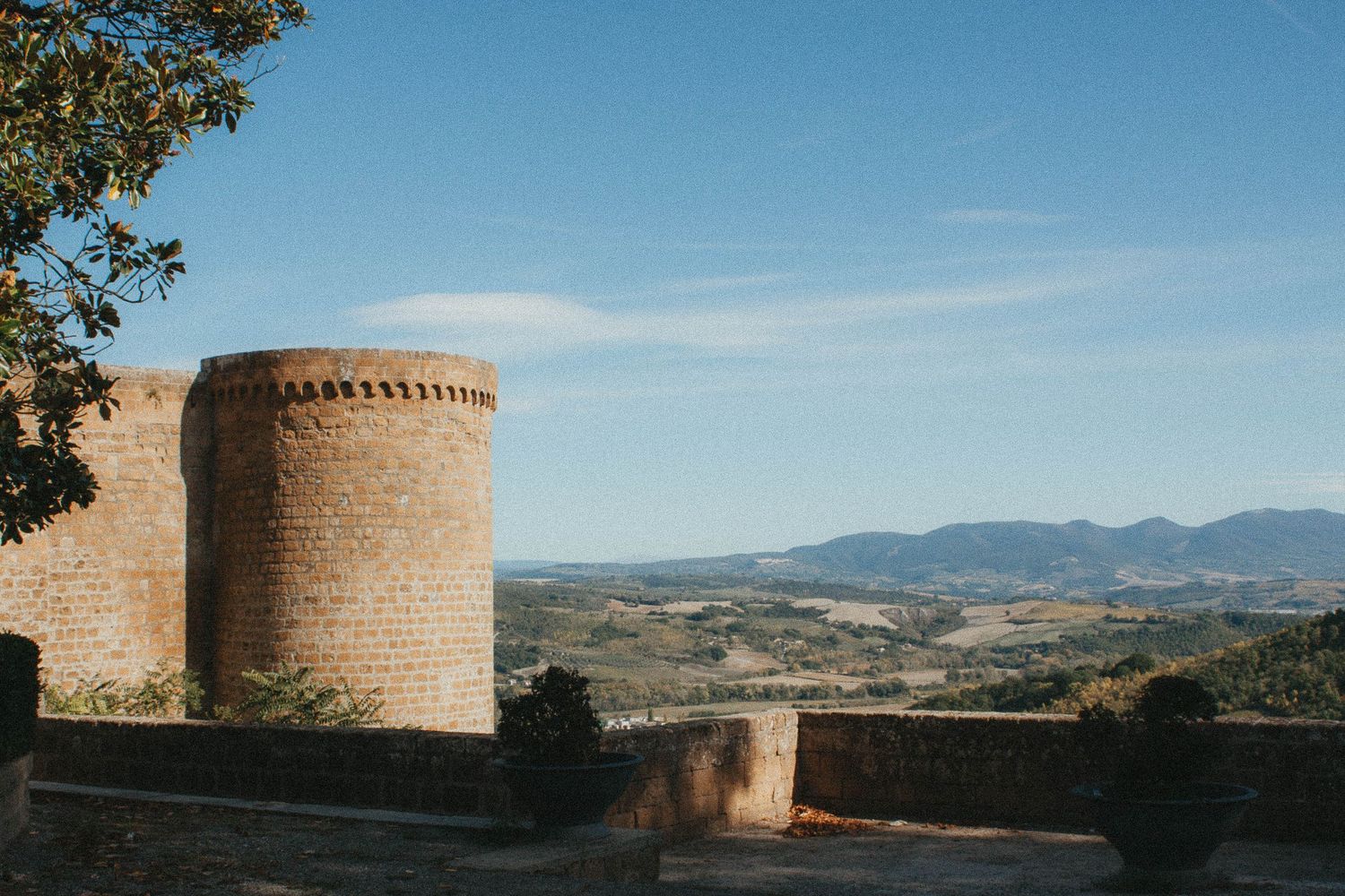 A medieval brick tower overlooking the sweeping Tuscan countryside under a bright blue sky.