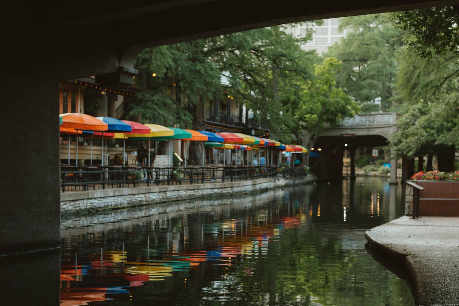 Colorful umbrellas reflect in the calm San Antonio River Walk canal beneath a bridge.