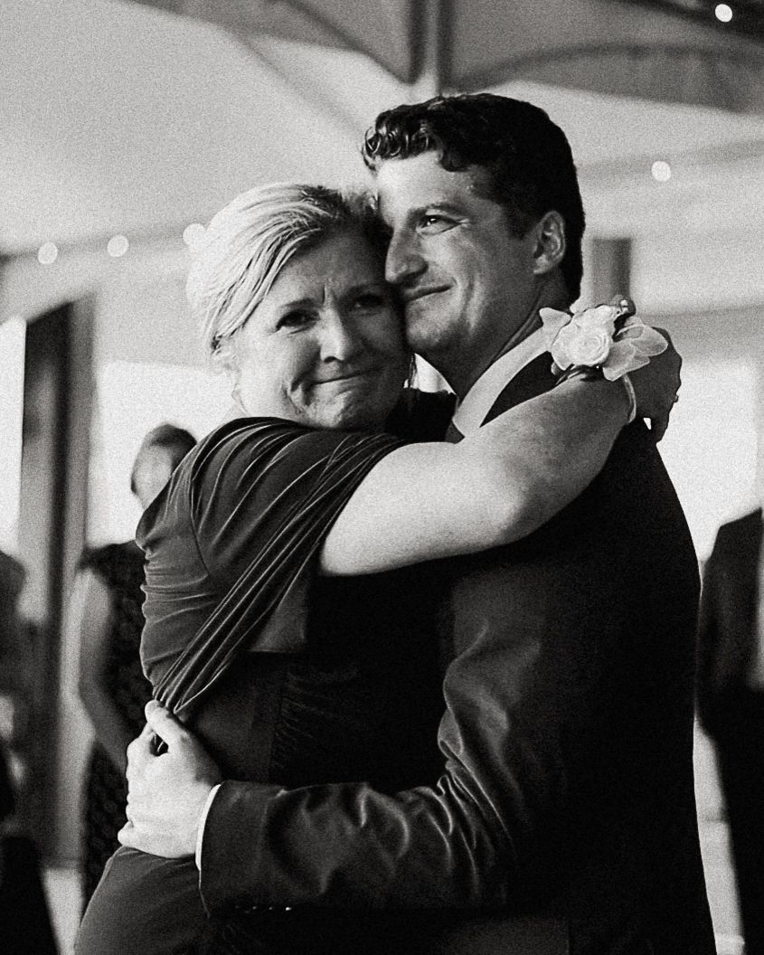 Black and white photo of a groom in a suit hugging an older woman, sharing a tender dance at a wedding reception.