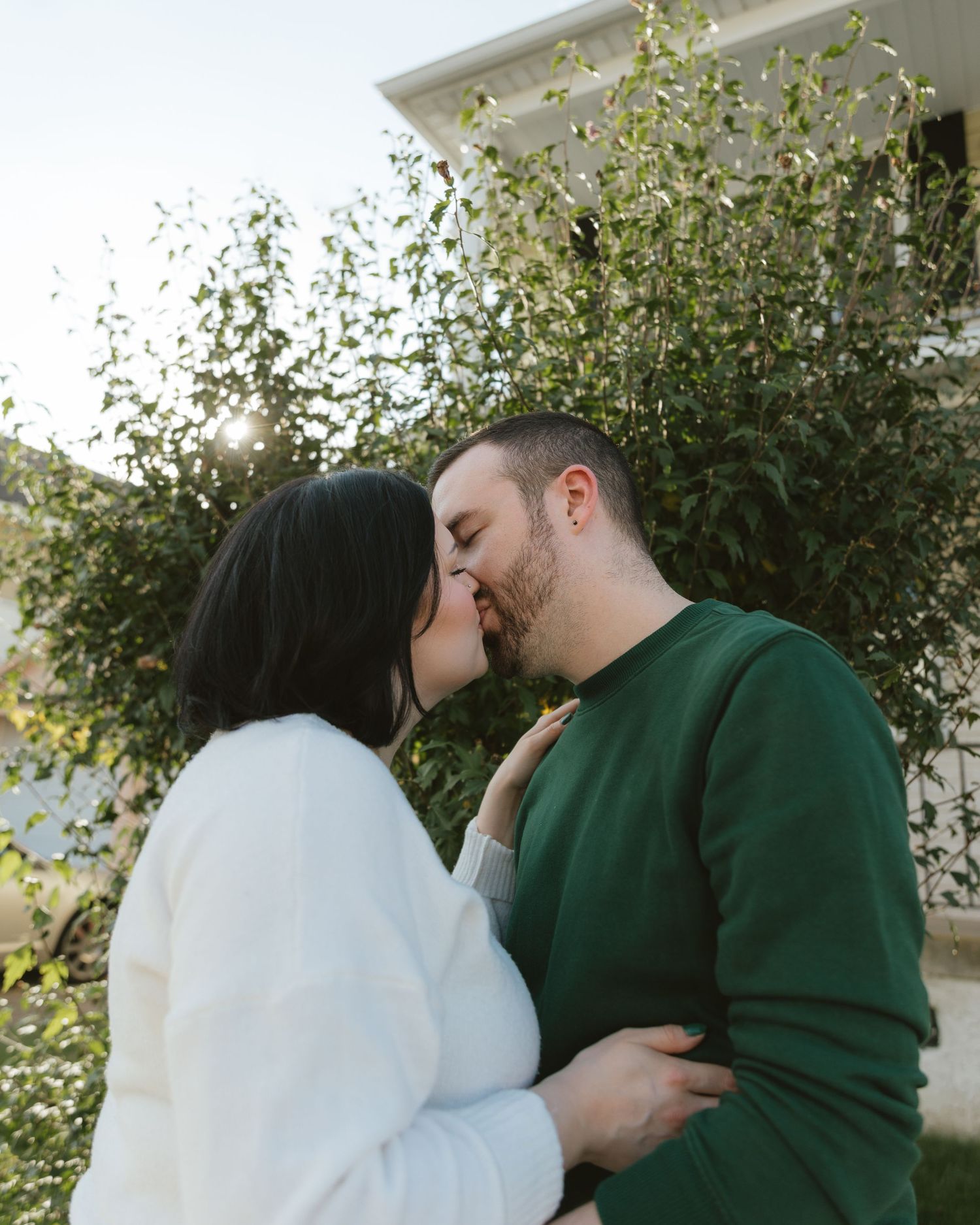A couple sharing a romantic kiss outdoors, surrounded by lush green foliage, wearing white and green sweaters.