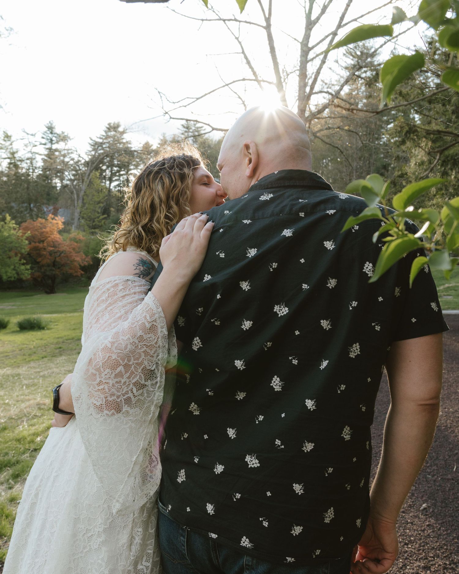 A couple shares a romantic kiss outdoors, woman in lace dress embracing bald man in black floral shirt.