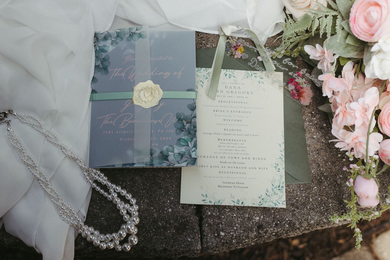 Elegant wedding invitation suite with floral design, green ribbon, wax seal, pearl necklace, and pink roses on stone surface.