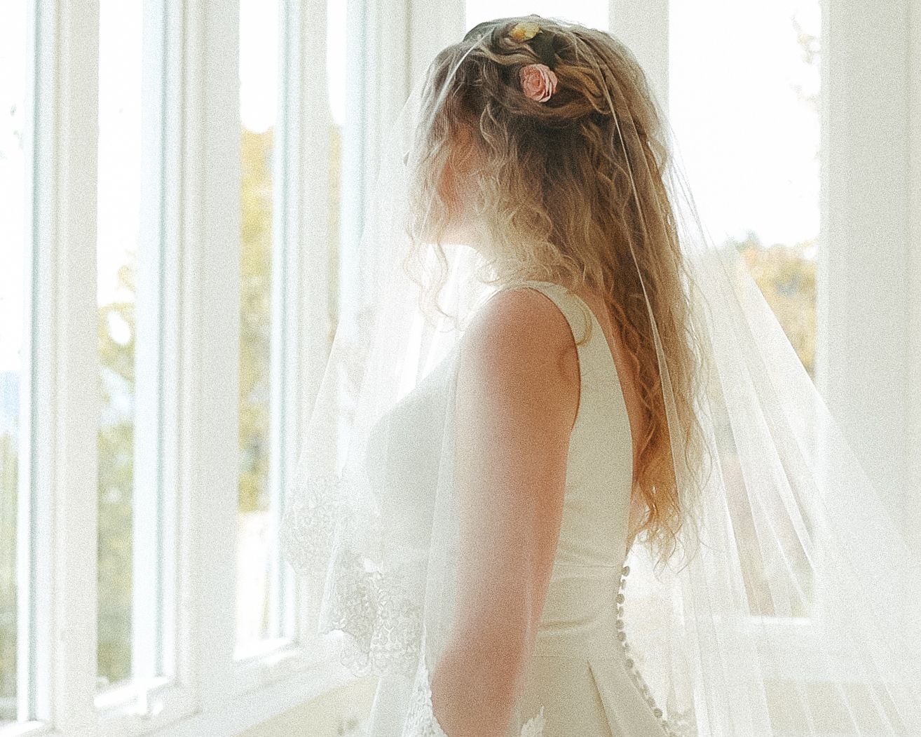 Bride wearing veil with floral hair accessories gazing out bright white window in wedding gown.