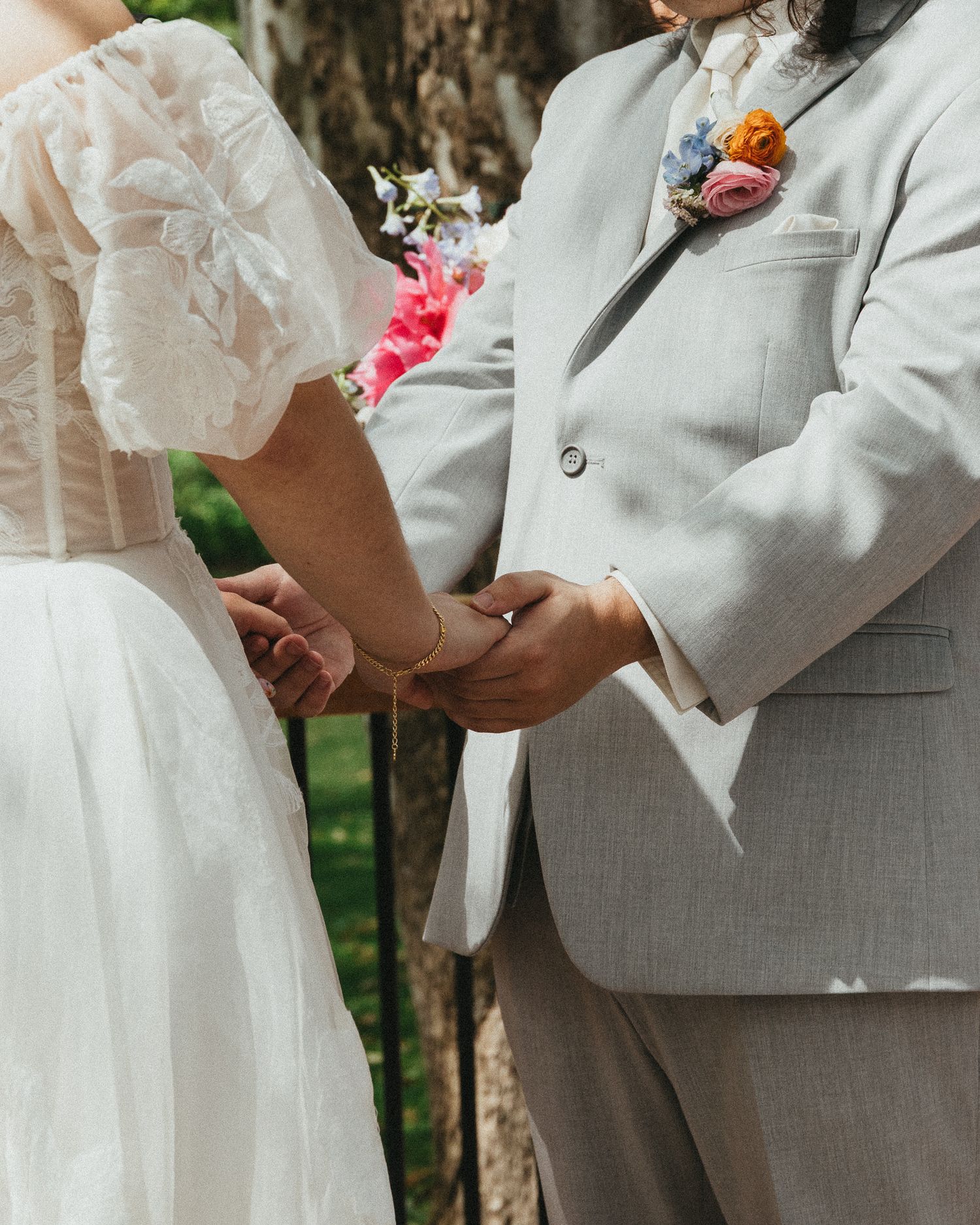 Bride in white lace dress and groom in gray suit holding hands during outdoor wedding ceremony.
