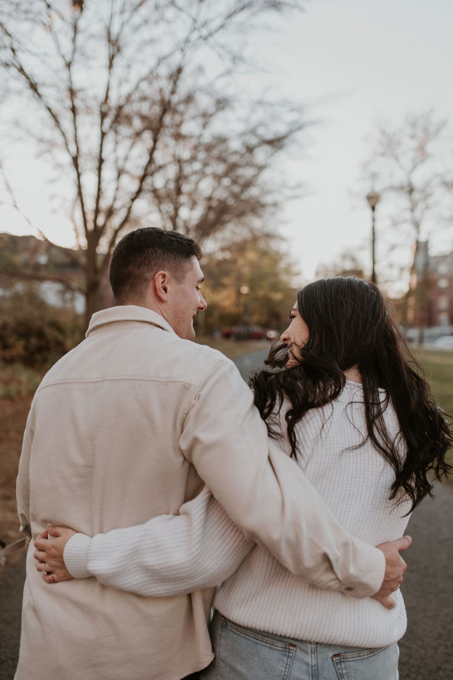 Couple embracing from behind on park path at dusk surrounded by autumn trees.