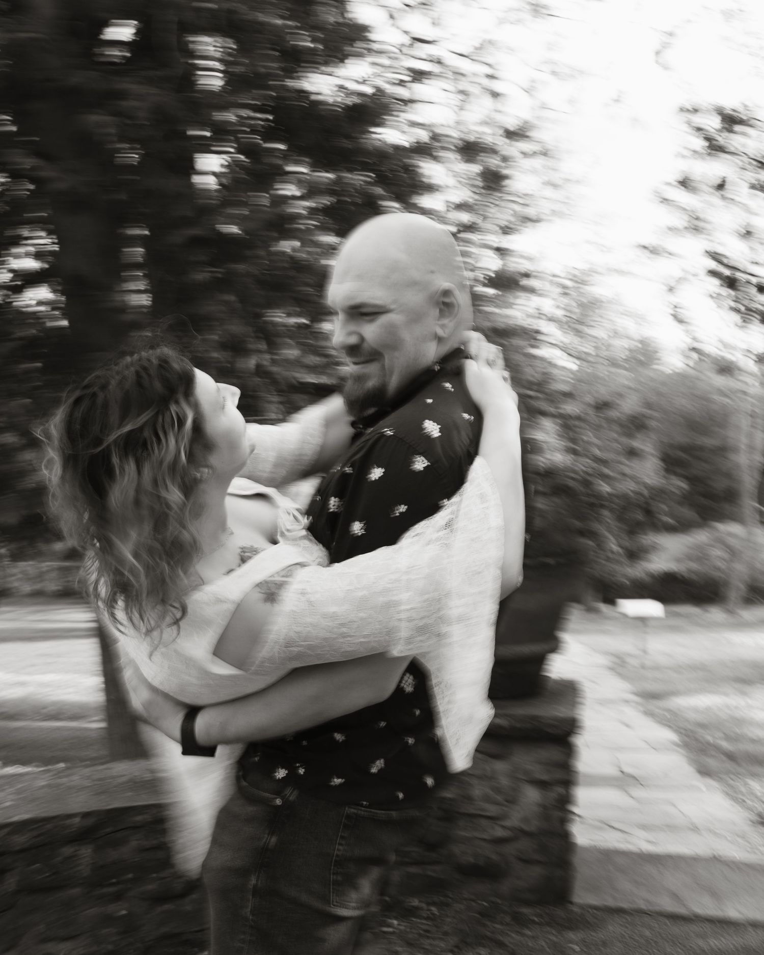 Black and white photo of a bald man and curly-haired woman embracing outdoors with trees in background.