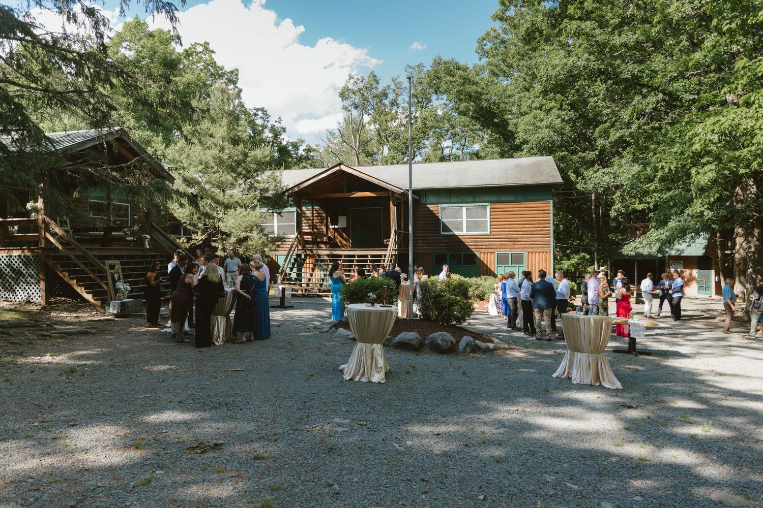 Guests mingle outdoors at a rustic woodland wedding venue surrounded by tall trees and log cabin buildings.