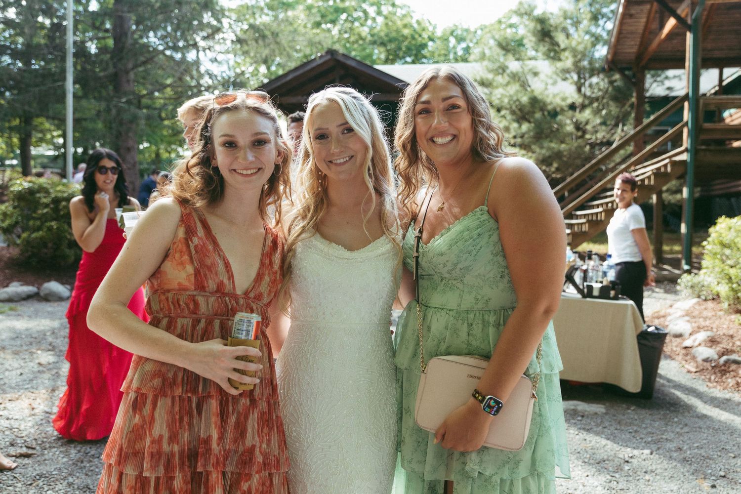 Three women in elegant dresses smile together outdoors at a wedding celebration surrounded by trees.